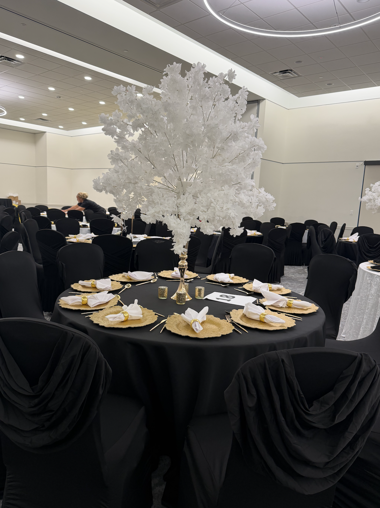 A banquet hall decorated with a round table featuring a tall white artificial tree centerpiece. The table is set with black tablecloths, gold placemats, white napkins with gold napkin rings, and small candles. Multiple chairs with black covers surrou