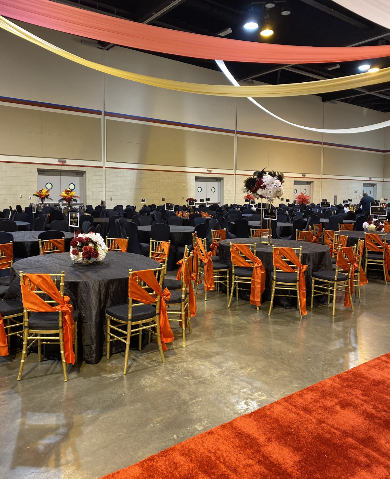 A banquet hall decorated with tables covered in black tablecloths, adorned with floral centerpieces, and gold chairs with orange sashes tied around them. Overhead, colorful drapes hang from the ceiling. The room has a polished concrete floor and a hi