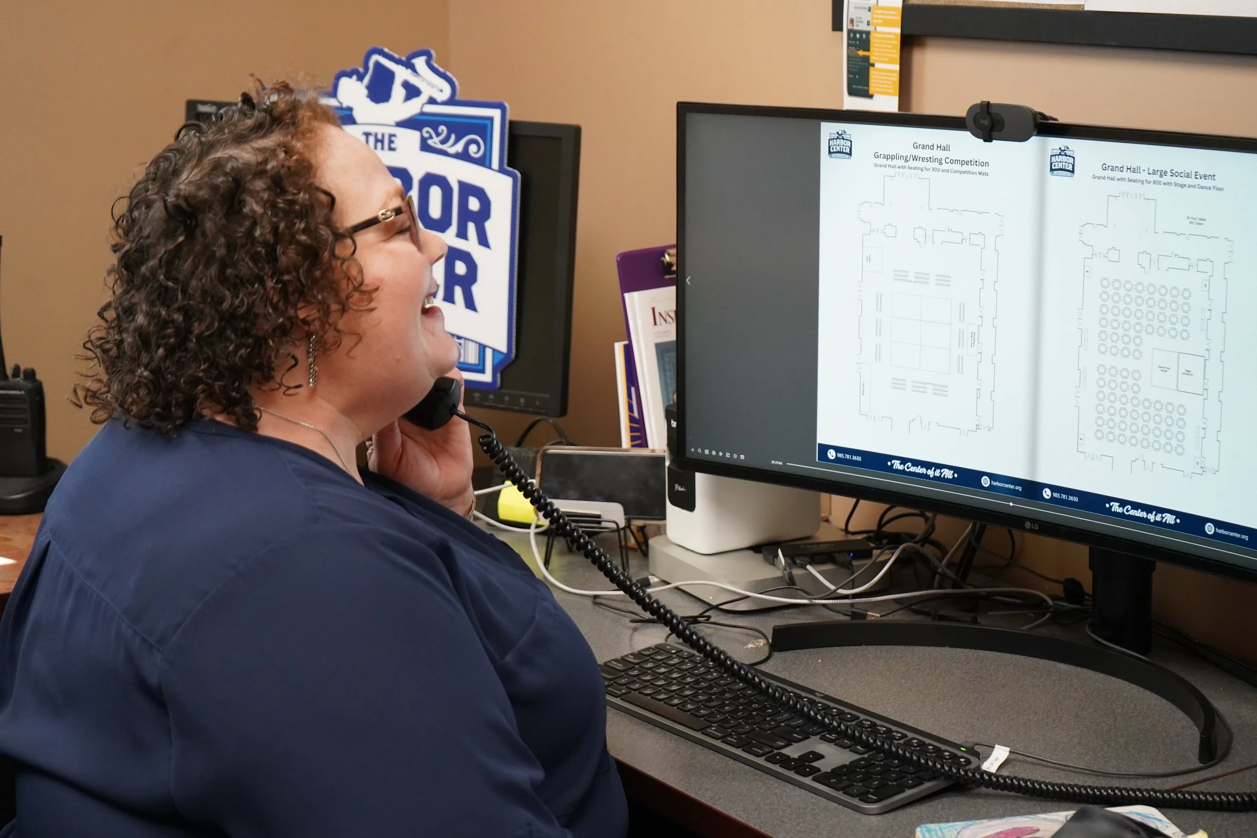 A woman with curly hair and glasses smiling while on a phone call at her desk, with a computer monitor displaying floor plans for an event space.