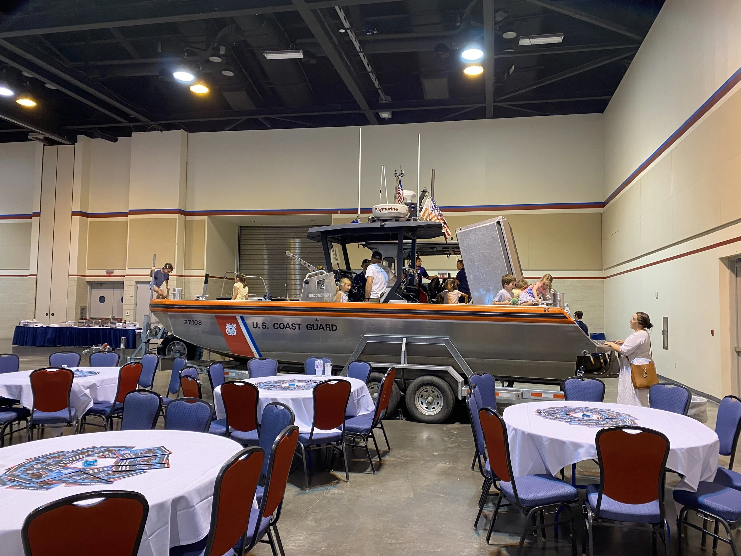 Indoor event with a U.S. Coast Guard boat on a trailer as a display. There are round tables with white tablecloths and chairs arranged around them in the foreground. Children and adults are around the boat, some are on the boat, and others are standi