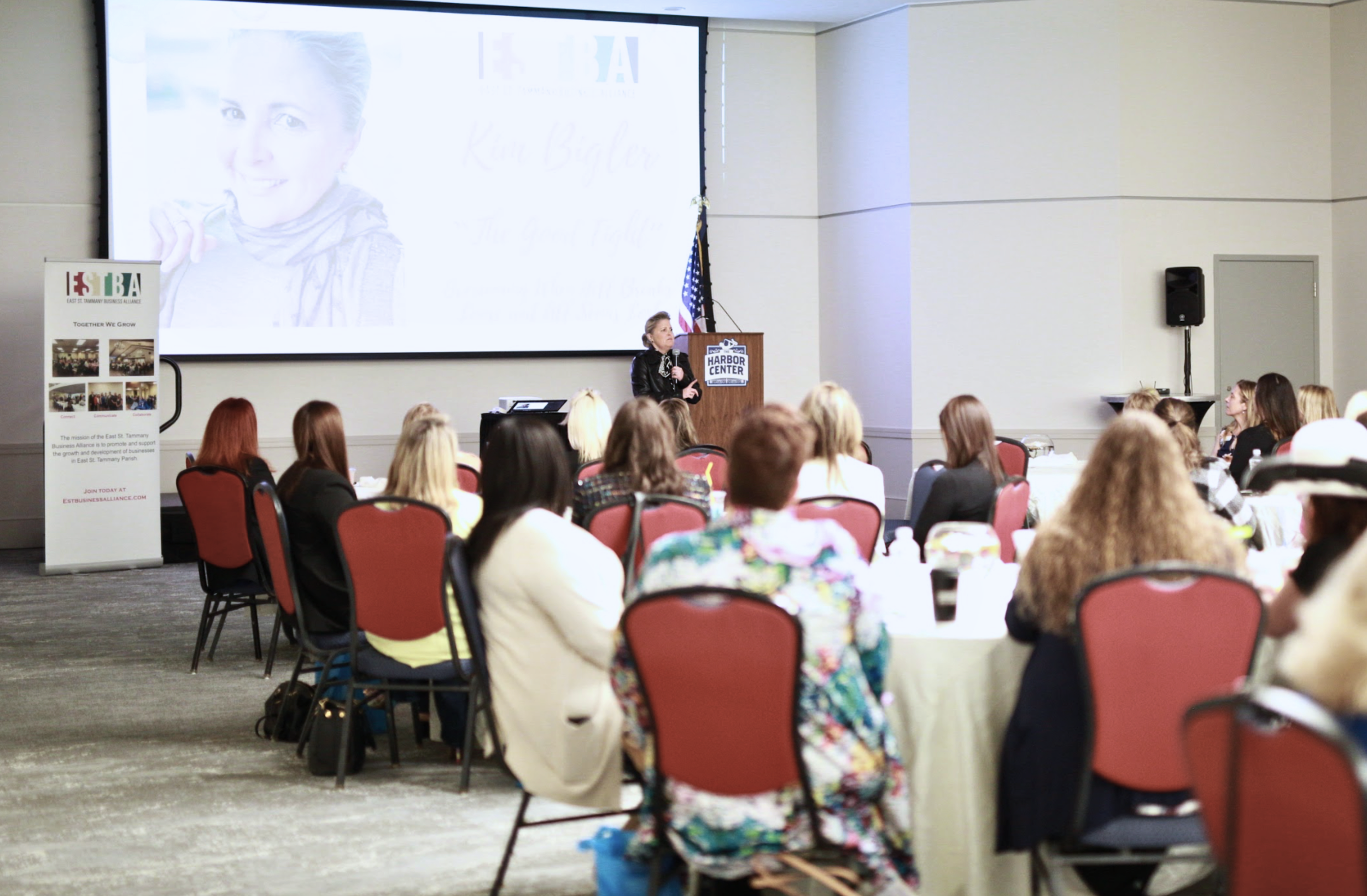 A woman is giving a presentation at a conference in a large room with round tables and red chairs. There is a large screen behind her showing her photo and some text. The audience is attentively listening to her.