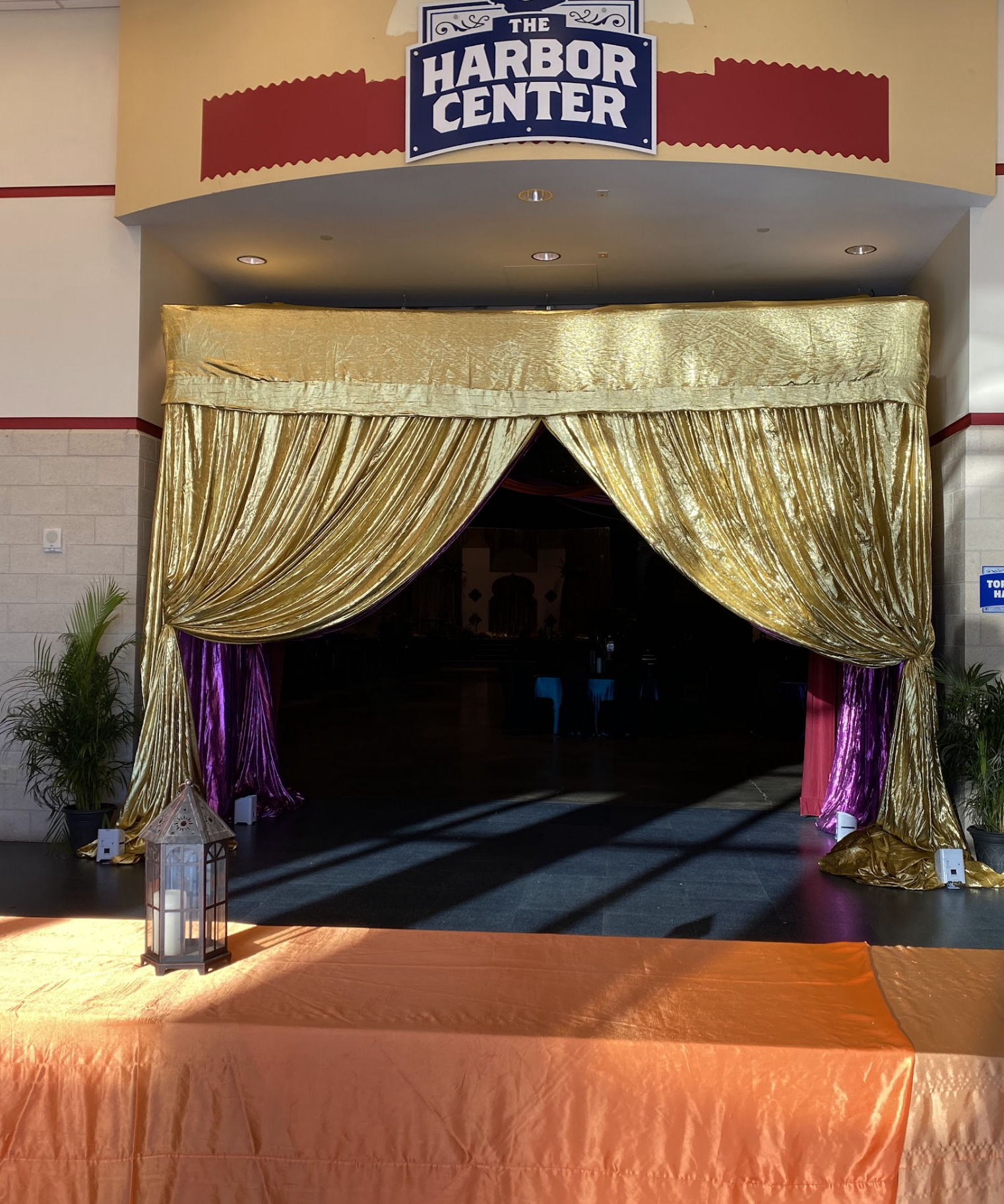 Decorative stage with gold curtains at The Harbor Center, with plants, a lantern, and an orange table in front.