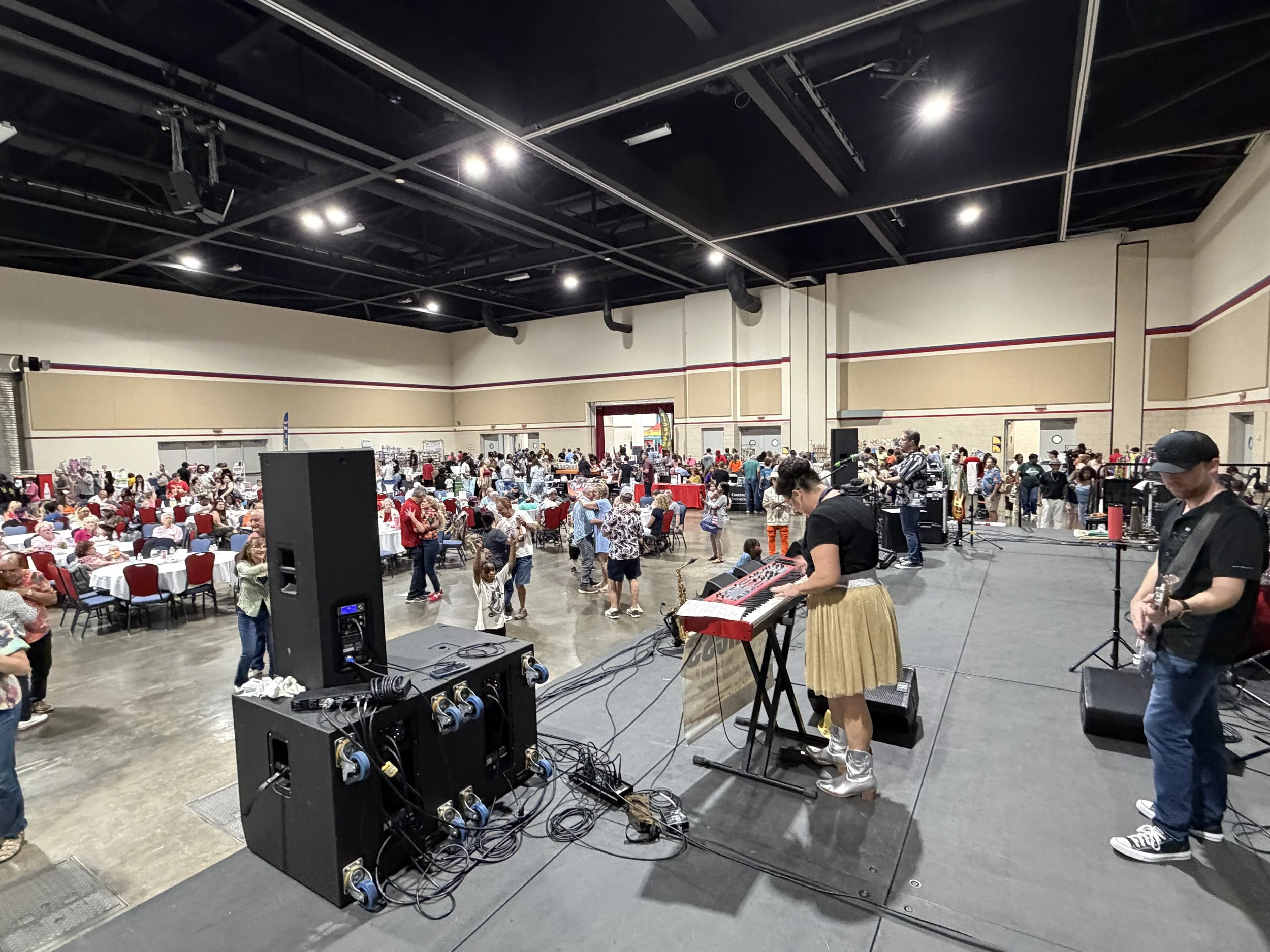 Musicians perform on stage at a large indoor event with many attendees seated at tables and some dancing in the background.