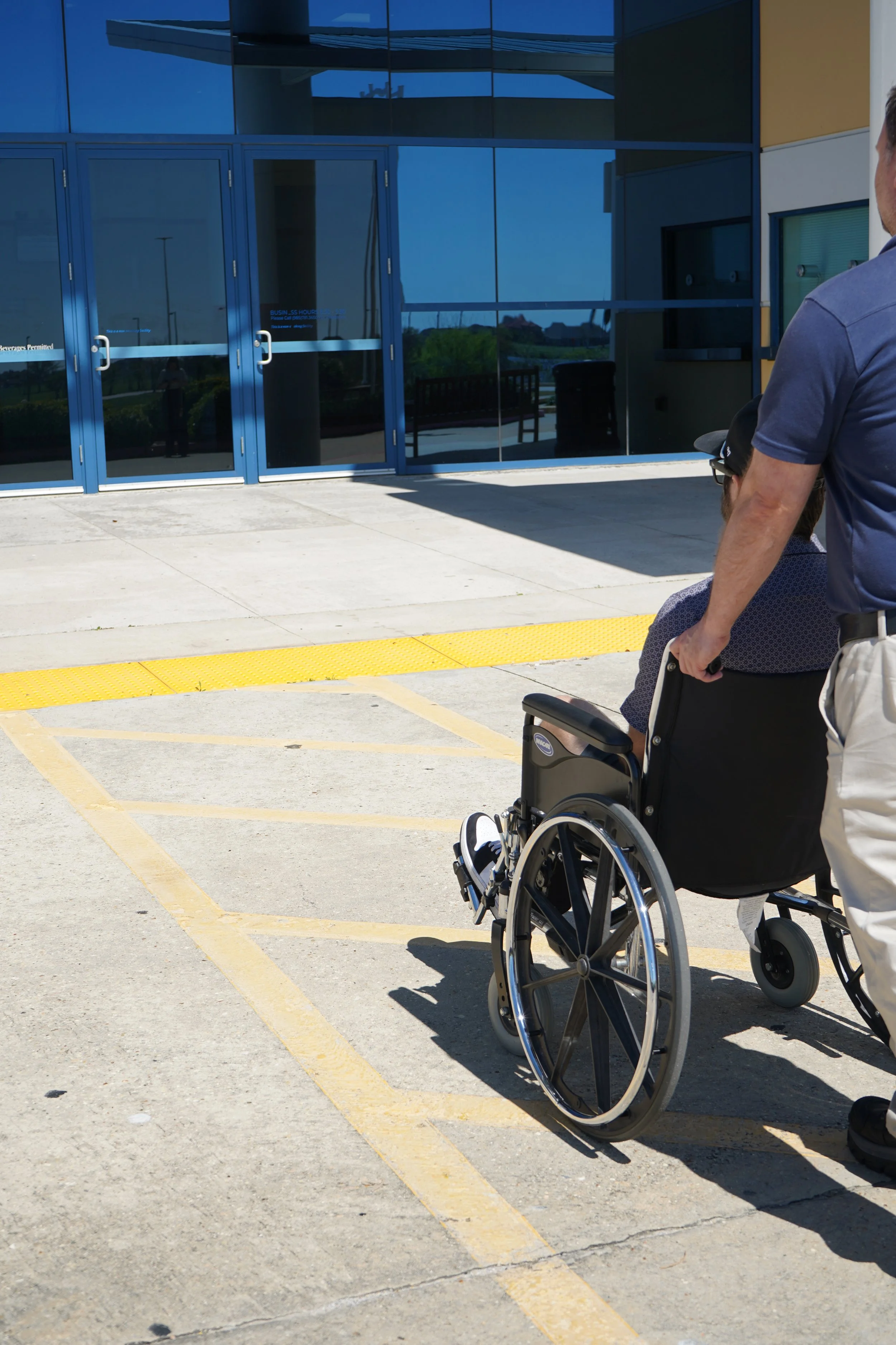 A person in a wheelchair being pushed outside a modern building with glass doors.