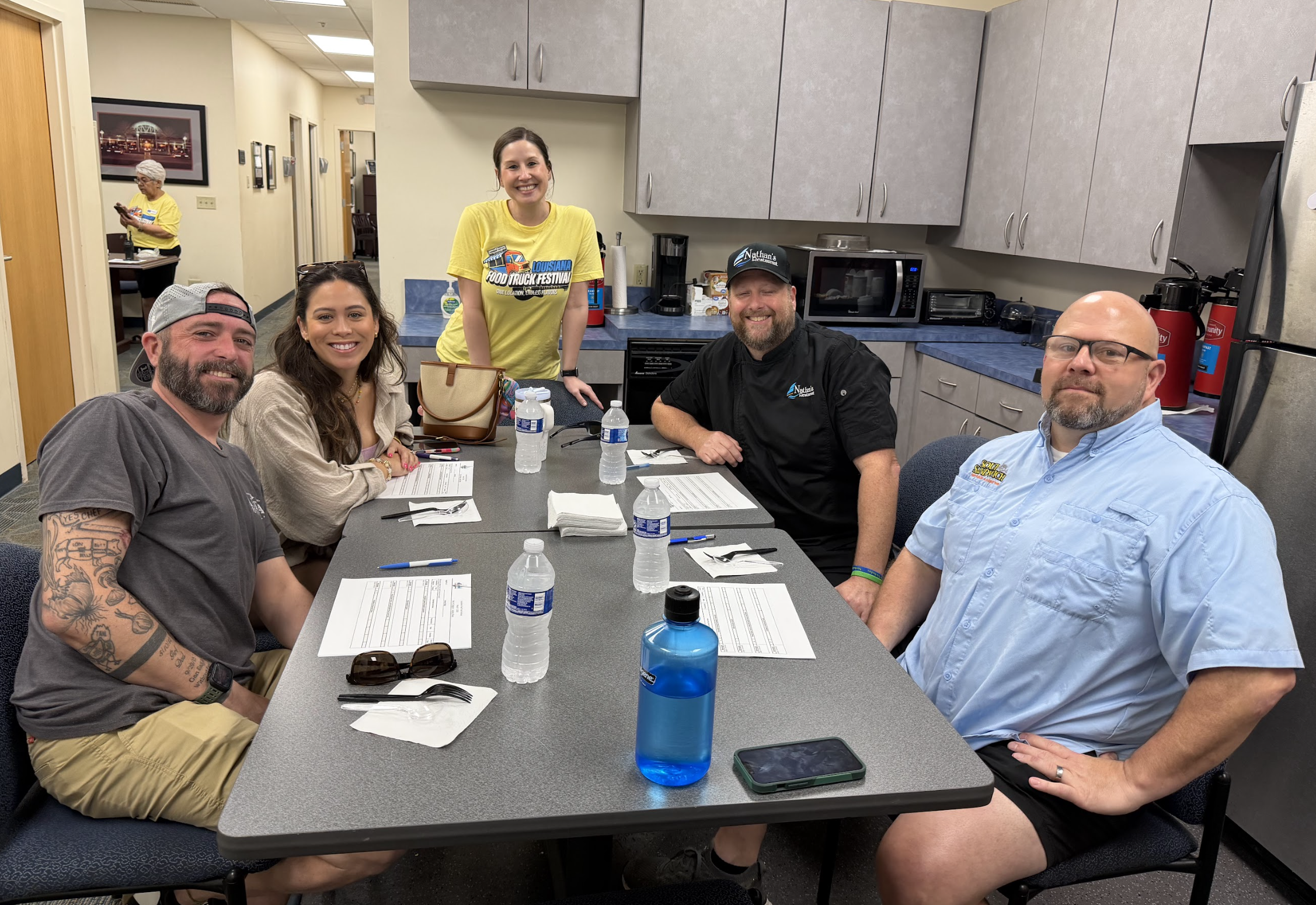 People sitting around a table with water bottles and papers, smiling for a photo in a kitchen-like room.
