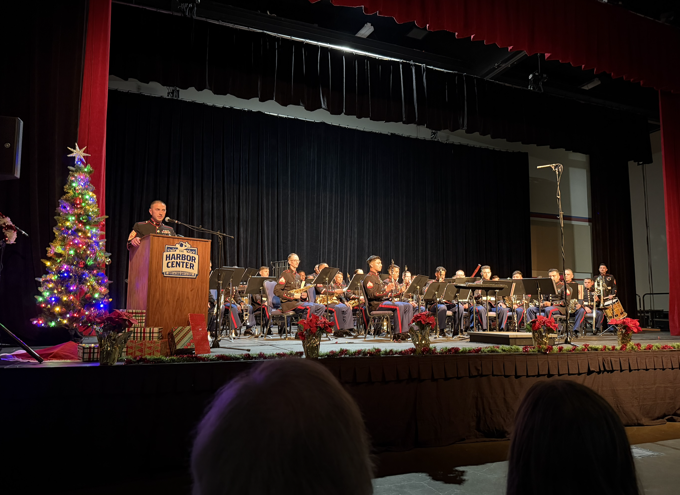 Military band performing on stage at Harbor Center decorated for Christmas with a lit Christmas tree, poinsettias, and holiday presents.