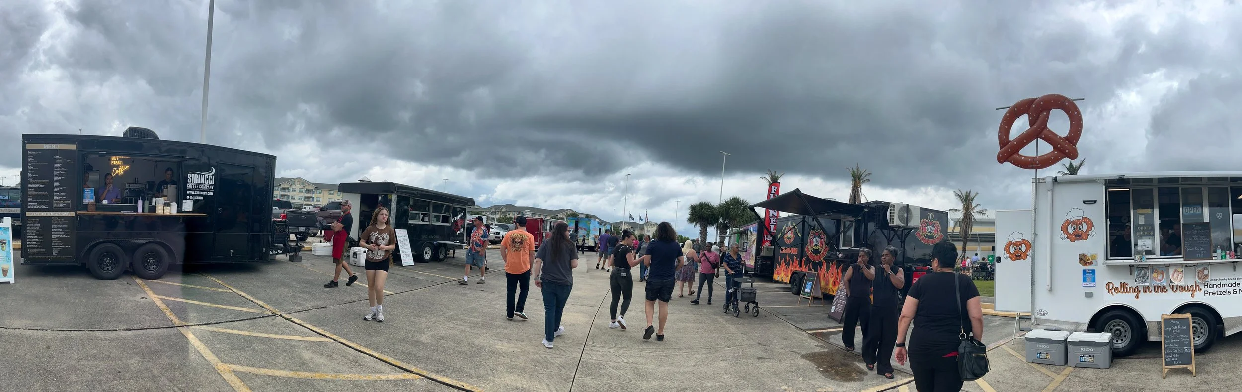 Food trucks parked in a lot on a cloudy day with people walking around and ordering food.