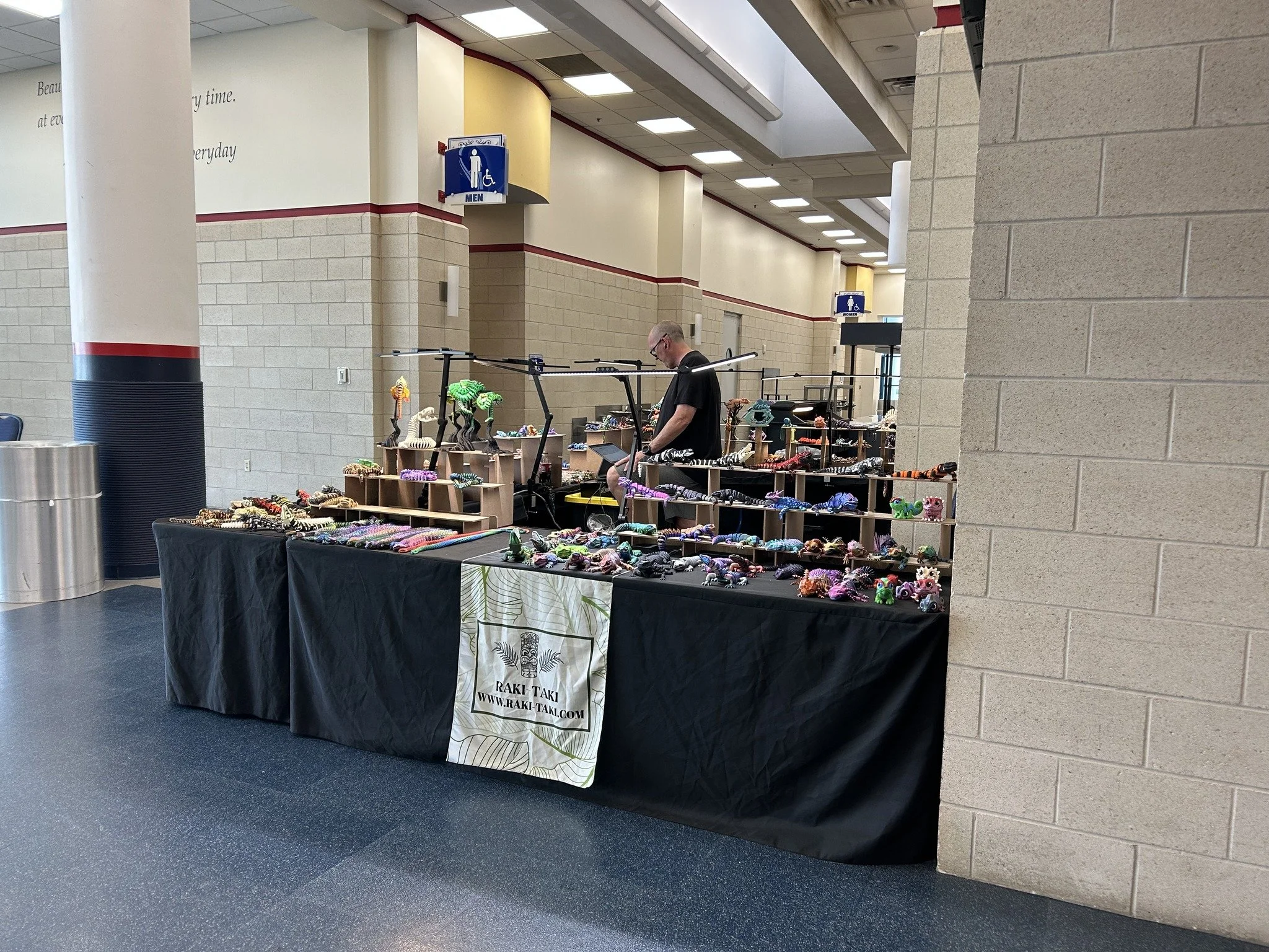 A vendor table displaying colorful handmade jewelry and crafts at an indoor market or event, with a person browsing the items.
