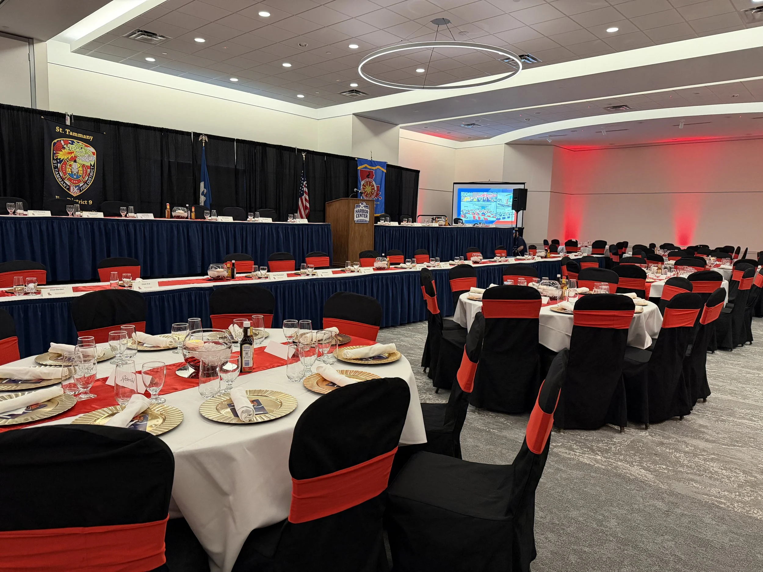 A large banquet room set up for a formal event, featuring round tables with black and red chairs, tables decorated with white tablecloths, glassware, and place settings, a long head table with a dark blue skirt, a podium, and a large screen in the ba