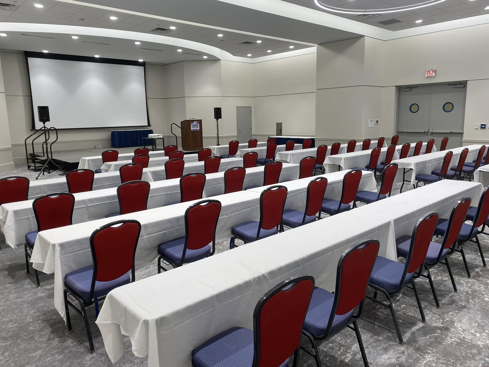 Empty conference room with rows of red and blue chairs, white tables, a large screen, and a podium at the front.
