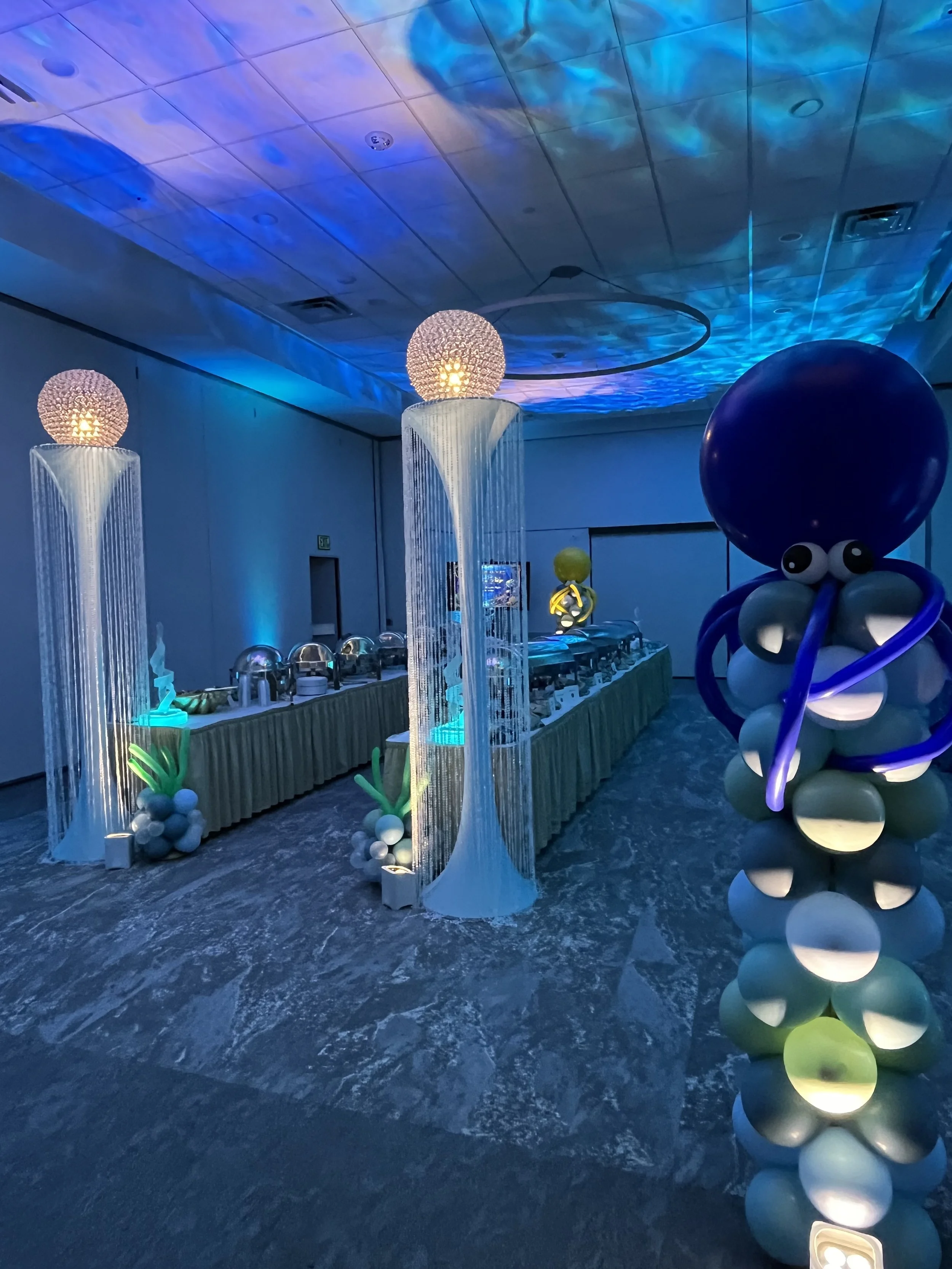 An indoor event space decorated with marine-themed balloons and illuminated fountain-like structures with orbs on top. The room has blue lighting and a buffet table in the background.