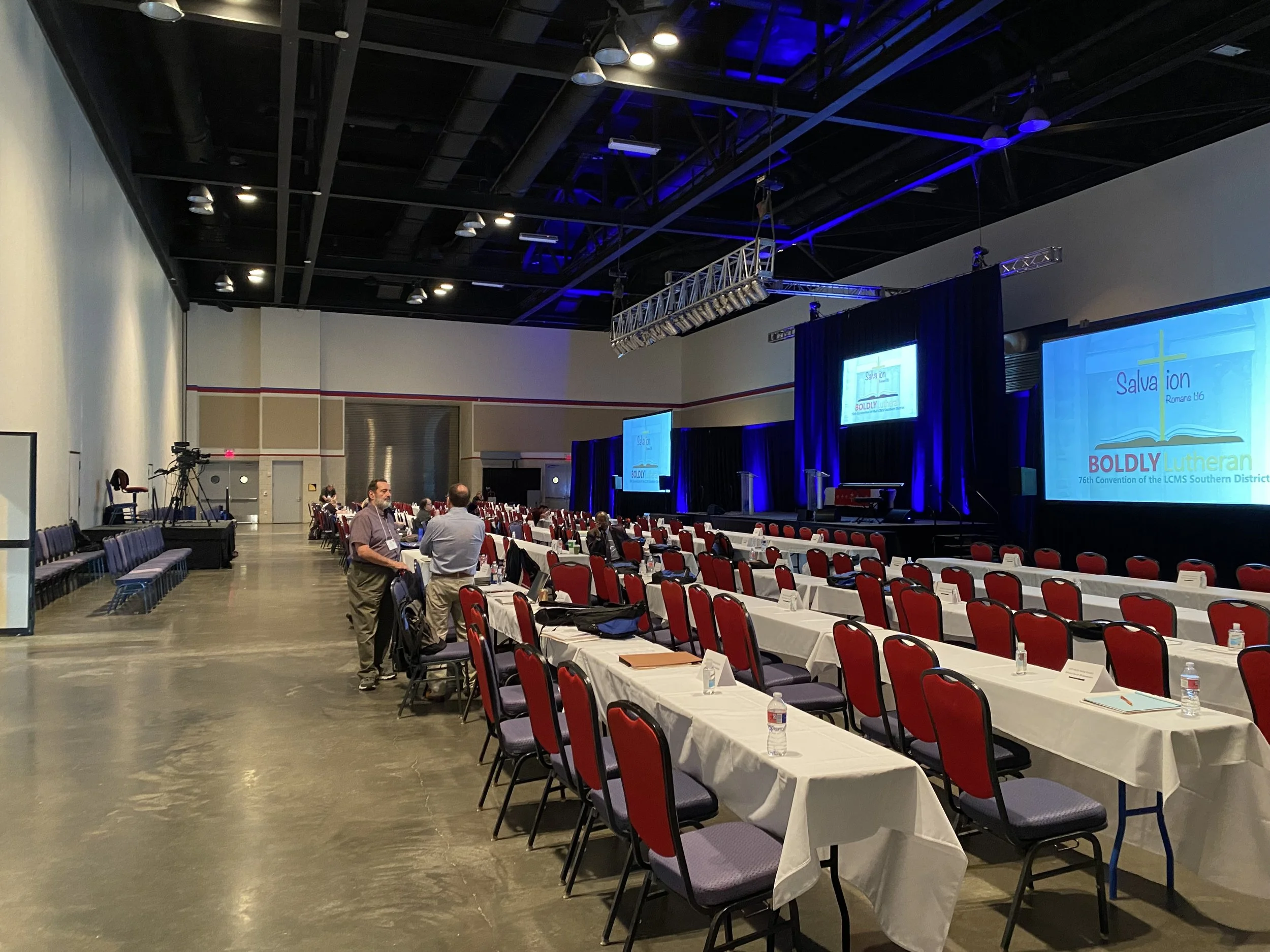 Conference room set up with rows of tables and red chairs, a stage with large screens displaying 'Salvation BOLDLY Lutheran' and event details, and some people standing and talking.