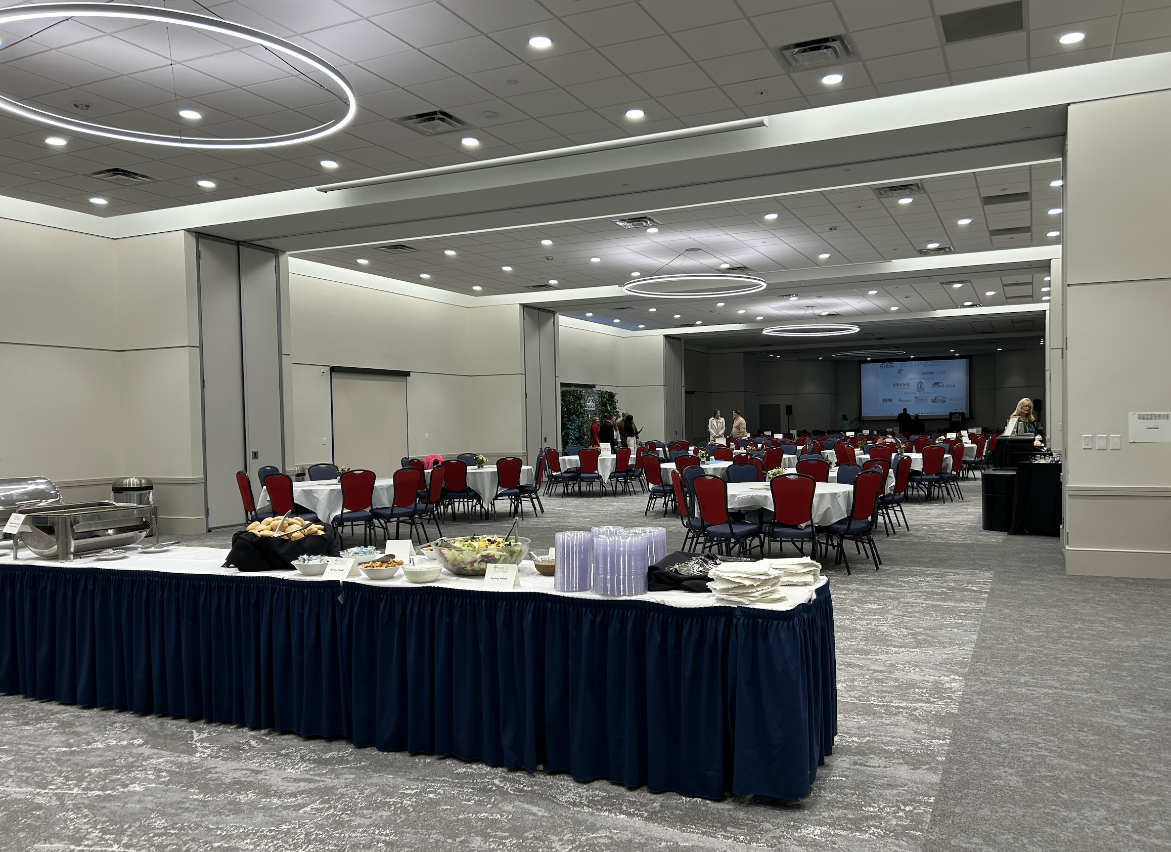 Empty banquet hall set up with round tables and red chairs, buffet table with food and utensils, and some people preparing.