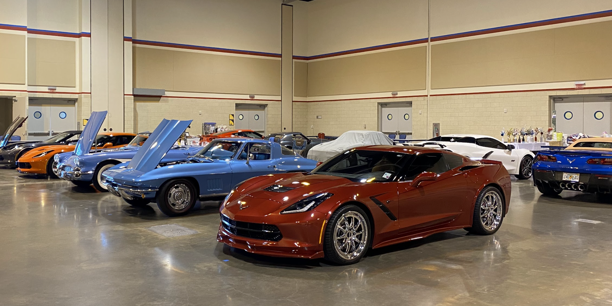 Multiple sports cars on display at an indoor car show, including a red Chevrolet Corvette in front, blue Corvettes with raised hoods, an orange Corvette, and other various cars with some covered and others with open hoods.
