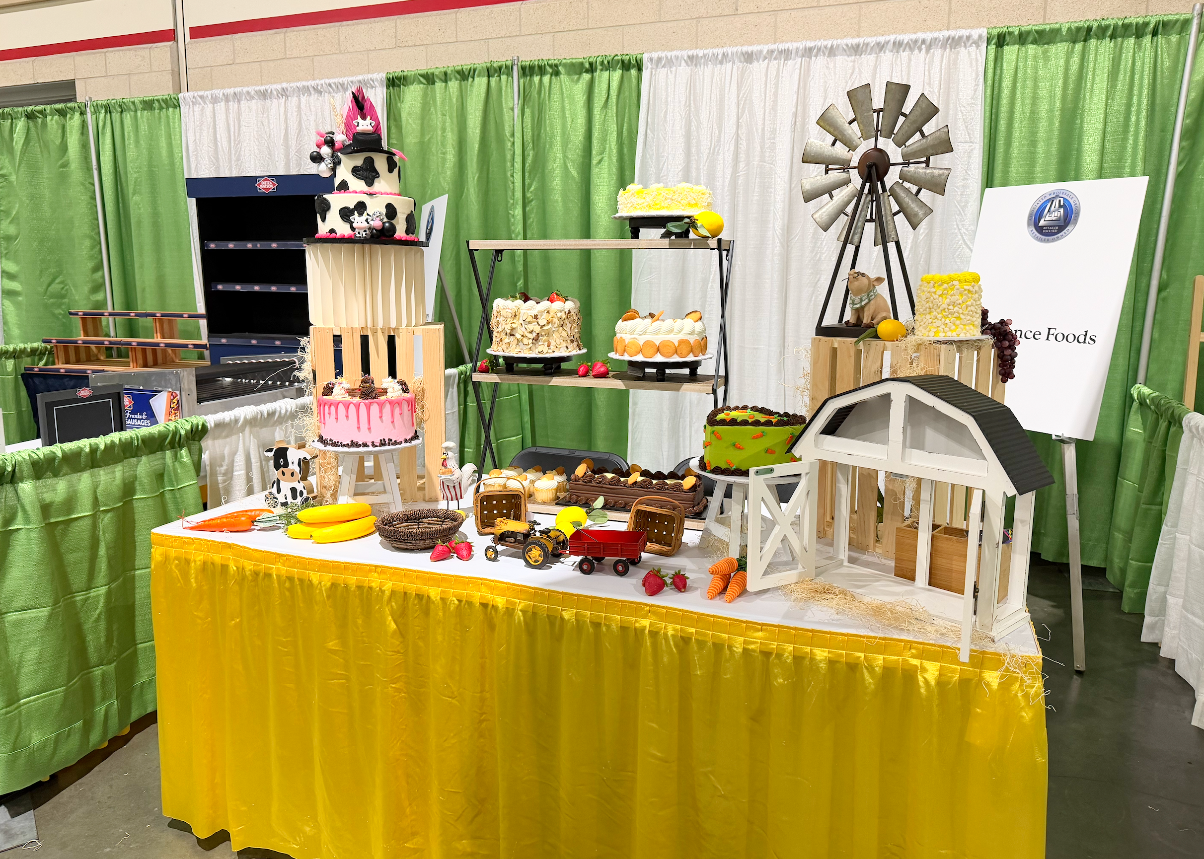Display of toy farm scene and decorated cakes at a cake show, with miniature barn, toy animals, and farm-themed desserts on a table covered with a yellow tablecloth, surrounded by green, white, and orange curtains.