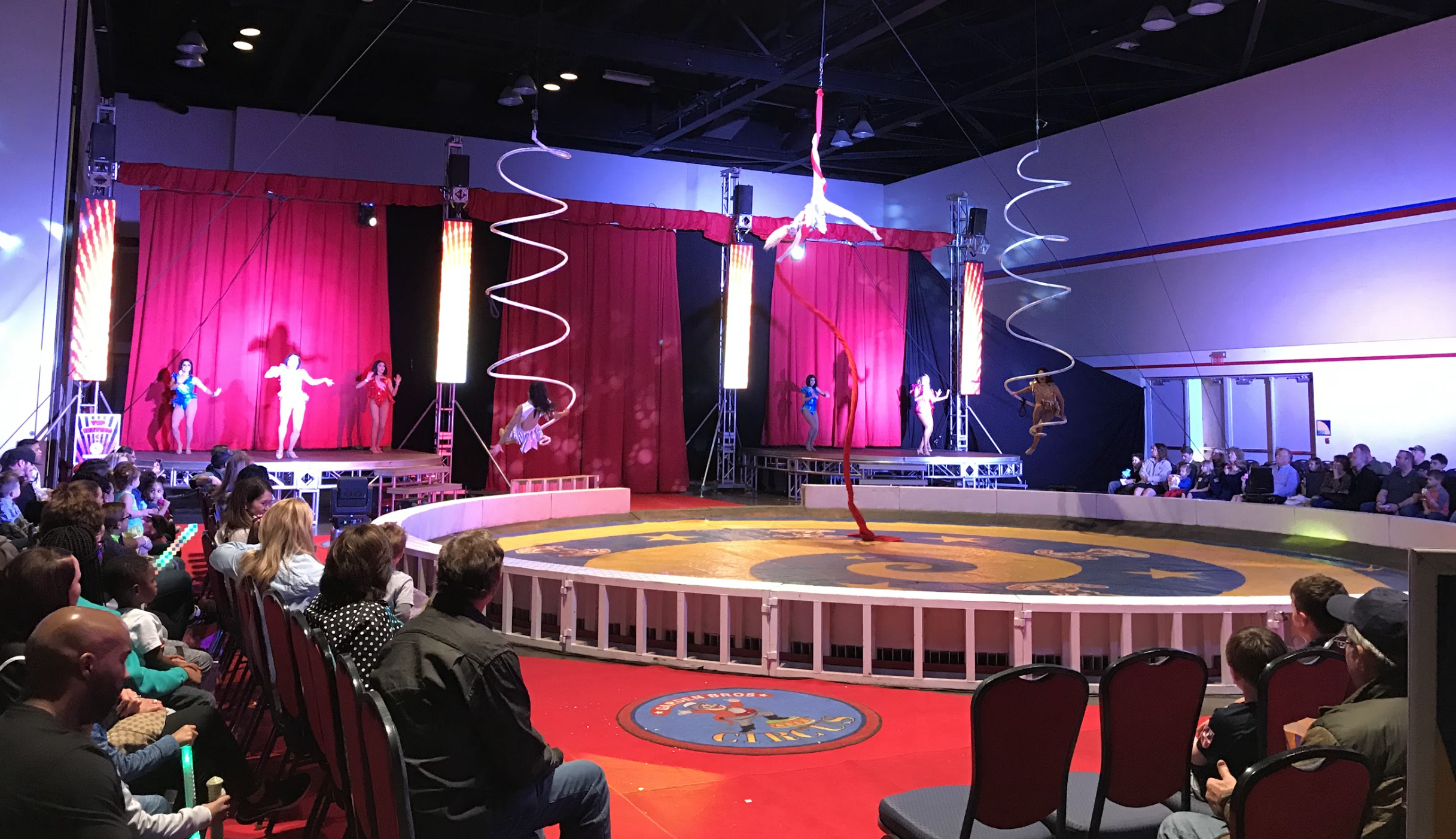 Circus performance with aerial acts and trapeze artists on stage, audience seated on red chairs, decorated with pink curtains and vertical LED screens.