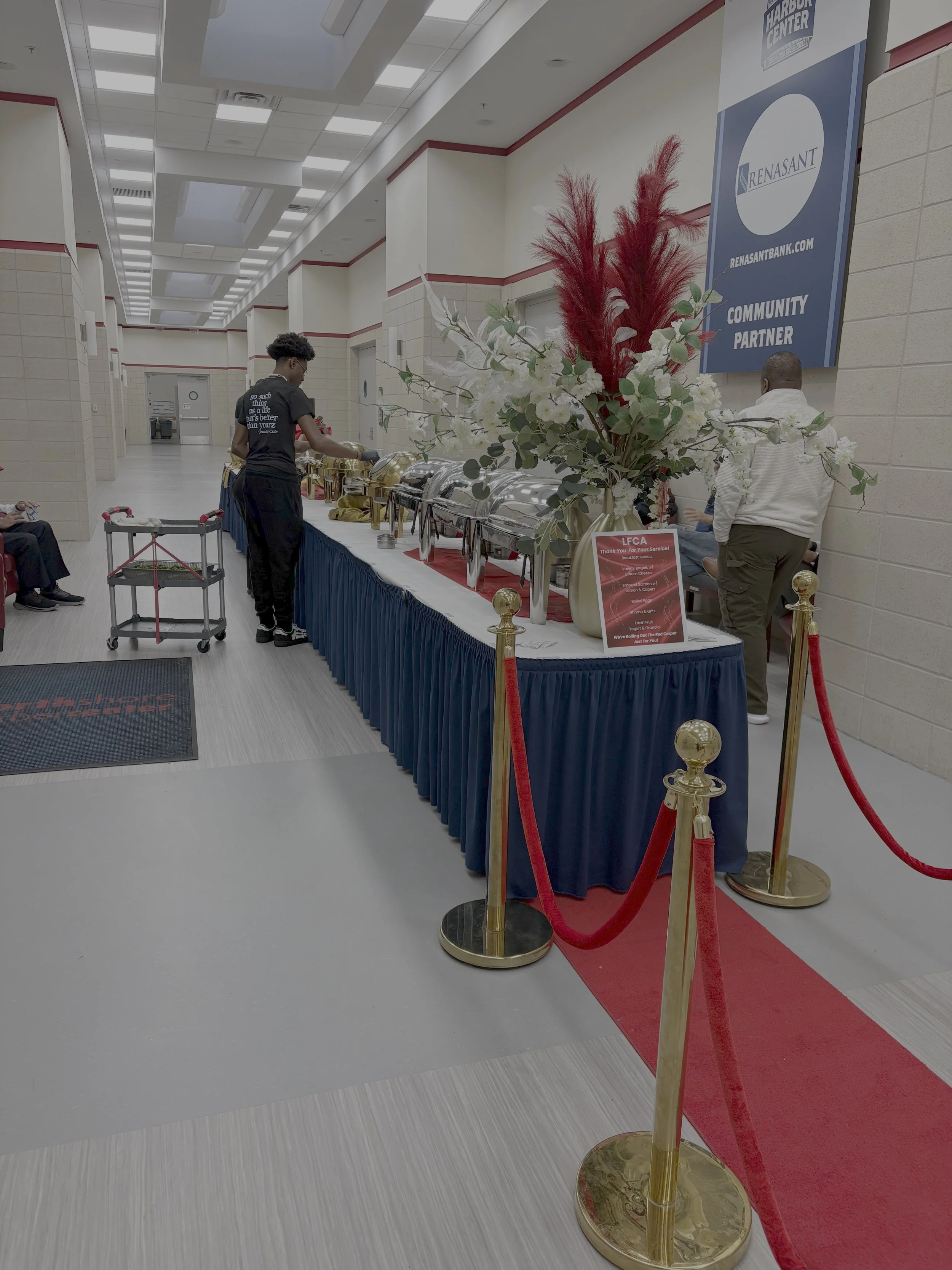 A buffet table with chafing dishes in a large, indoor event space, decorated with a large floral arrangement including white flowers, green leaves, and tall red feathers. Velvet ropes create a queue area, and there are people standing nearby. The roo