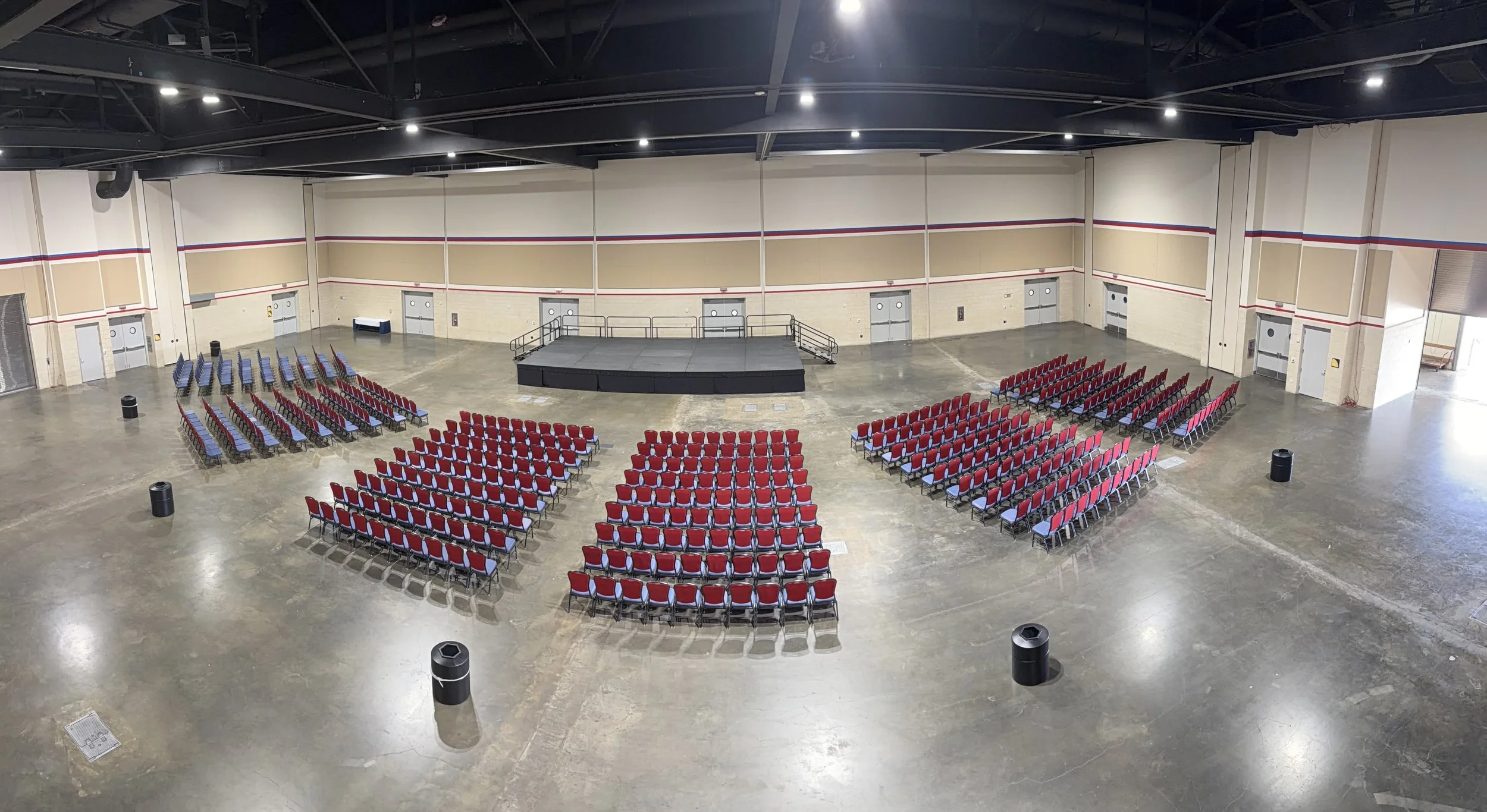 A large indoor event space with a stage in the center, surrounded by rows of chairs with red and blue upholstery. The room has a high ceiling with exposed black beams and lighting fixtures, beige walls with red and blue stripes, and a polished concre