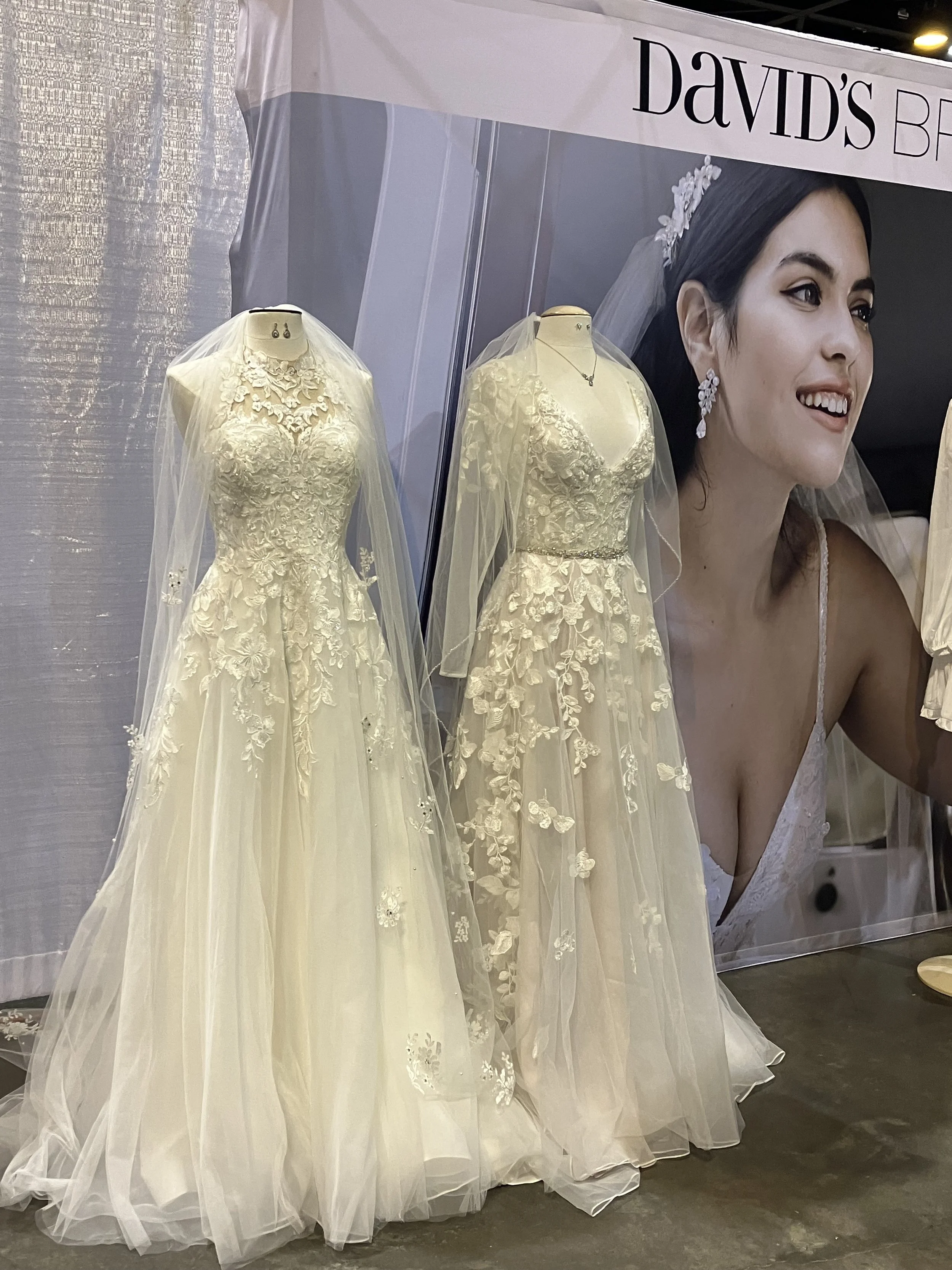 Two wedding dresses displayed on mannequins with veils and intricate lace details, positioned in front of a promotional poster featuring a bride.