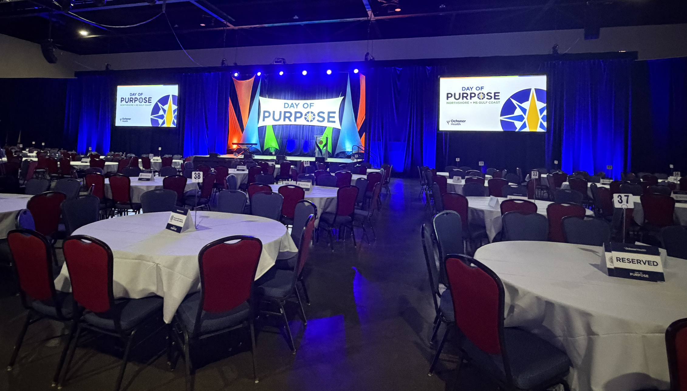 Empty banquet hall with round tables covered in white tablecloths, red and black chairs, and a stage with two large screens displaying a logo and text. The screens read 'Day of Purpose,' with colorful stage lighting.