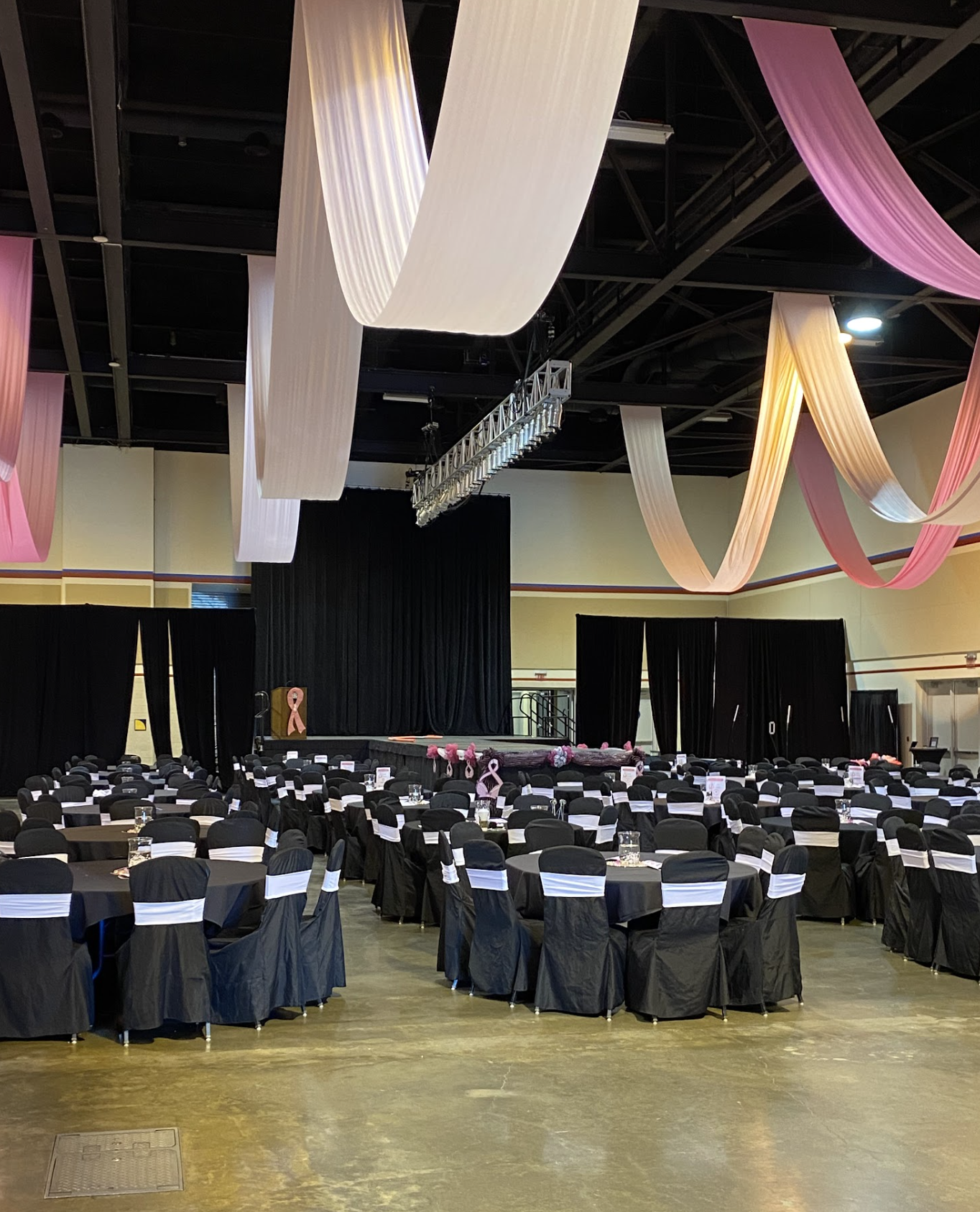 Event hall decorated with pink and white fabric drapes hanging from the ceiling, black tables and chairs with white sashes, a stage with a black curtain, pink ribbons, and breast cancer awareness symbols.