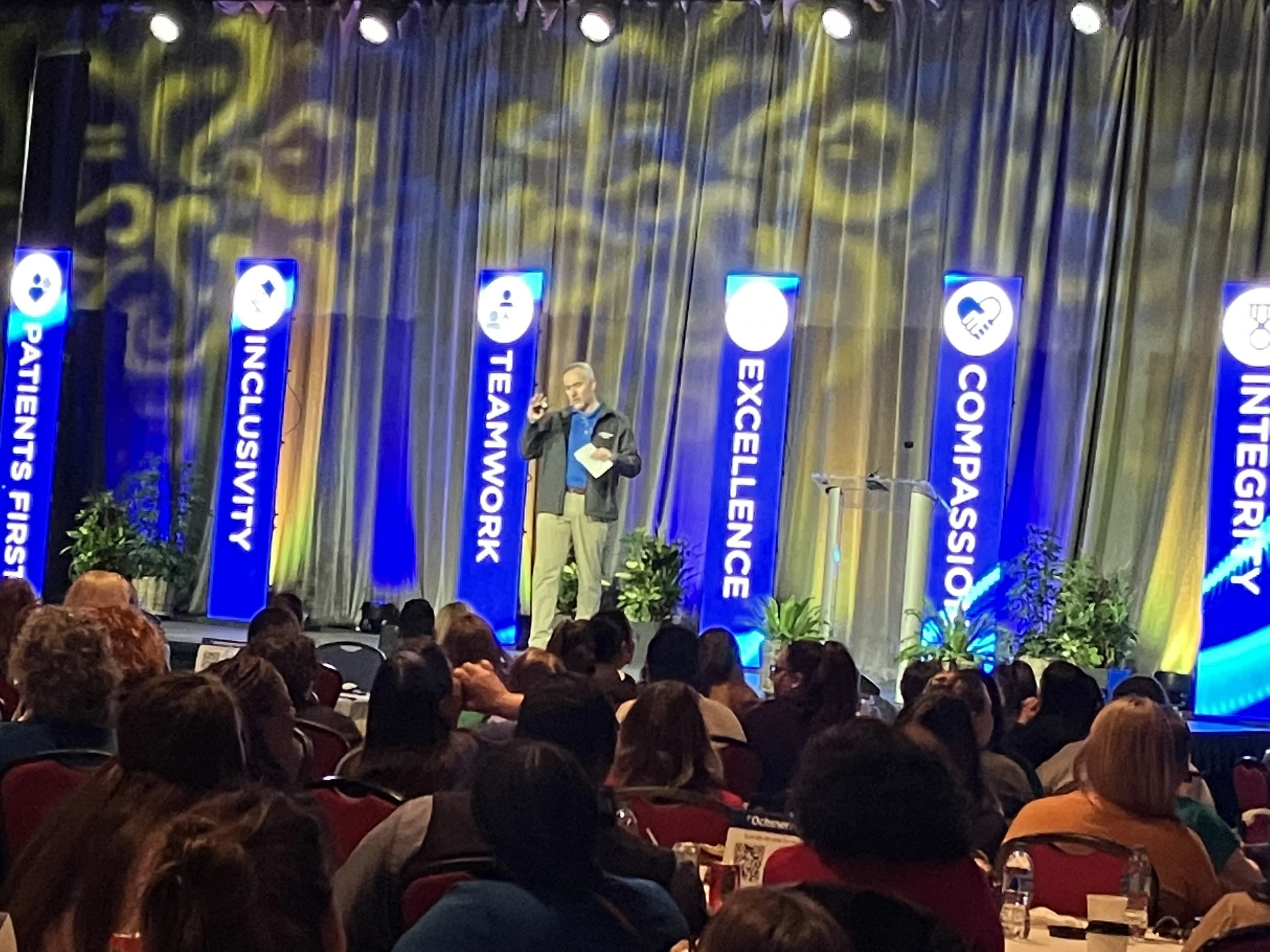 A person giving a presentation on stage at a conference with banners displaying words such as 'Patience First,' 'Inclusivity,' 'Teamwork,' 'Excellence,' 'Compassion,' and 'Integrity.' The audience is seated and watching.