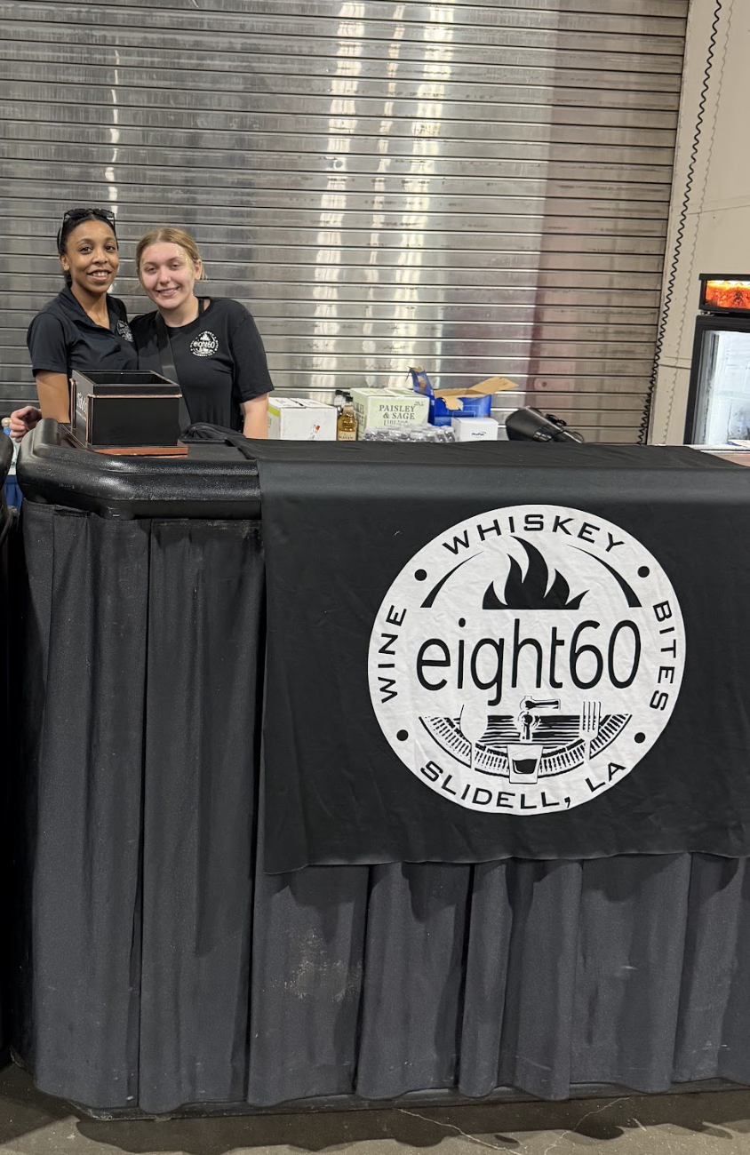 Two women smiling behind a bar with a black tablecloth displaying the eight60 logo, at Slidell, Louisiana, serving whiskey, wine, and bites.