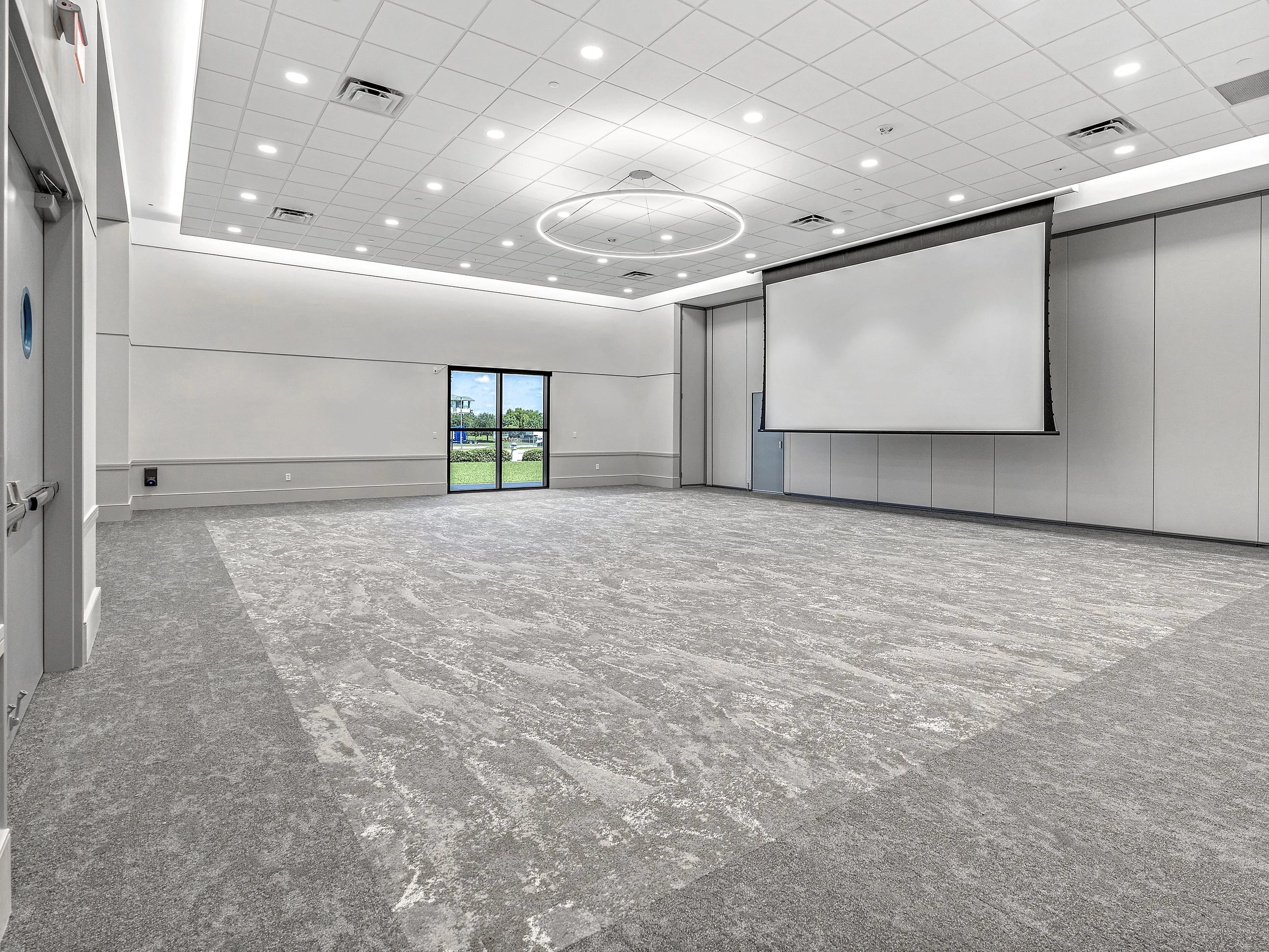 Empty conference room with a large projection screen, window showing green outdoors, and ceiling lights.
