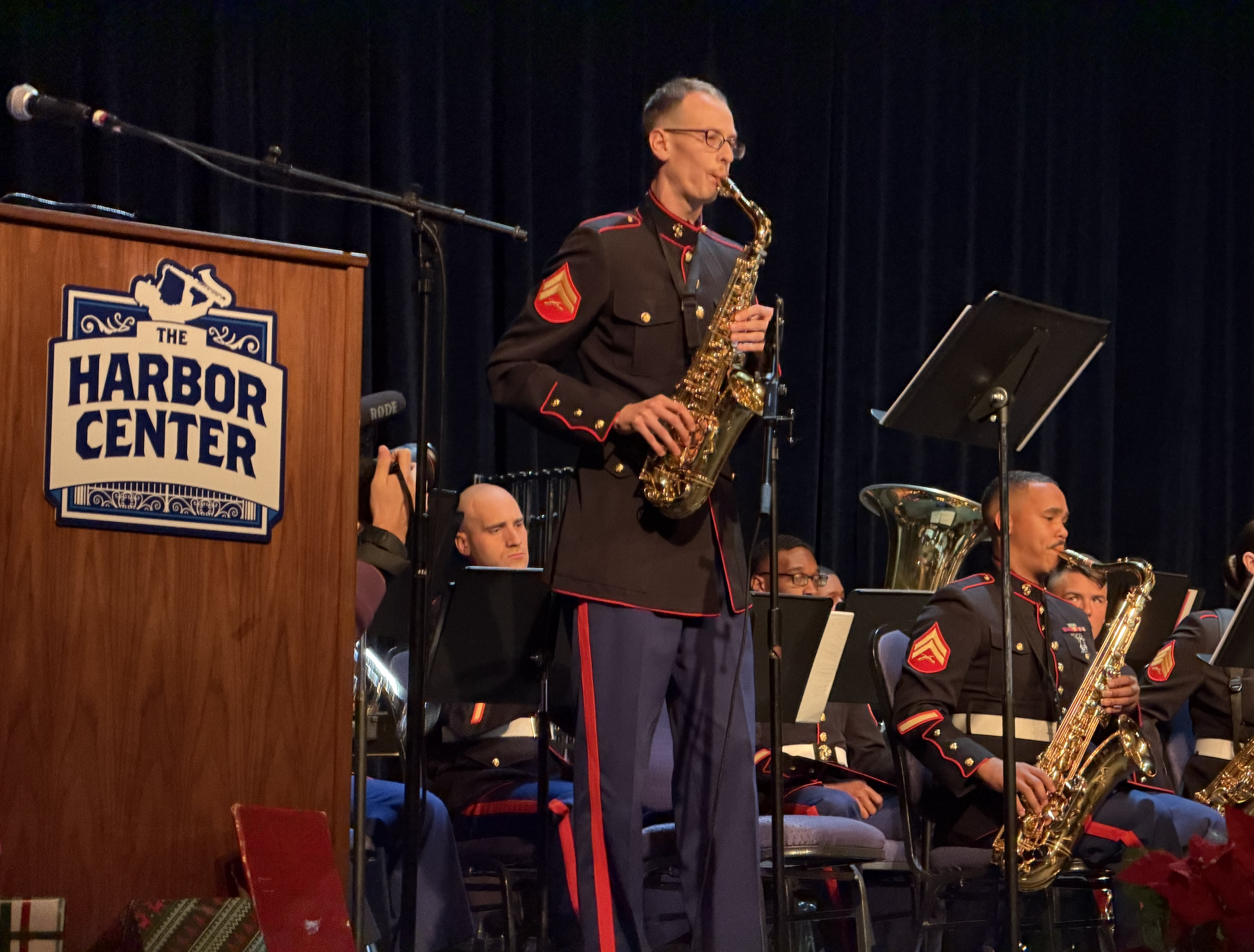 A military band performing on stage at the Harbor Center, with a saxophone player in the foreground and other musicians playing various instruments. The stage has a dark curtain backdrop and a wooden podium with the Harbor Center logo.