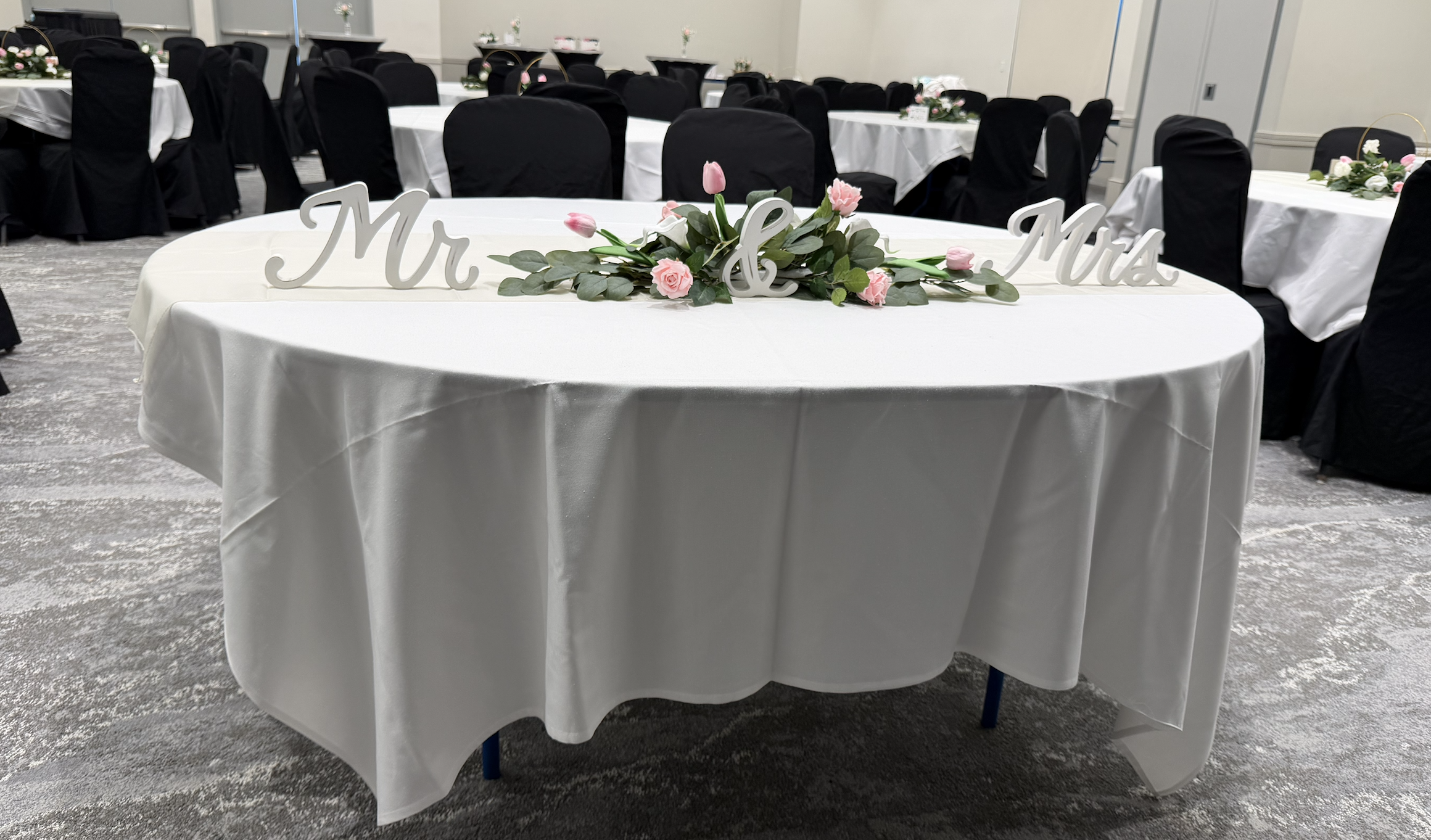 Wedding reception table with white tablecloth, floral centerpiece, and wooden 'Mr & Mrs' signs, in a banquet hall with black chairs covered in black cloth.