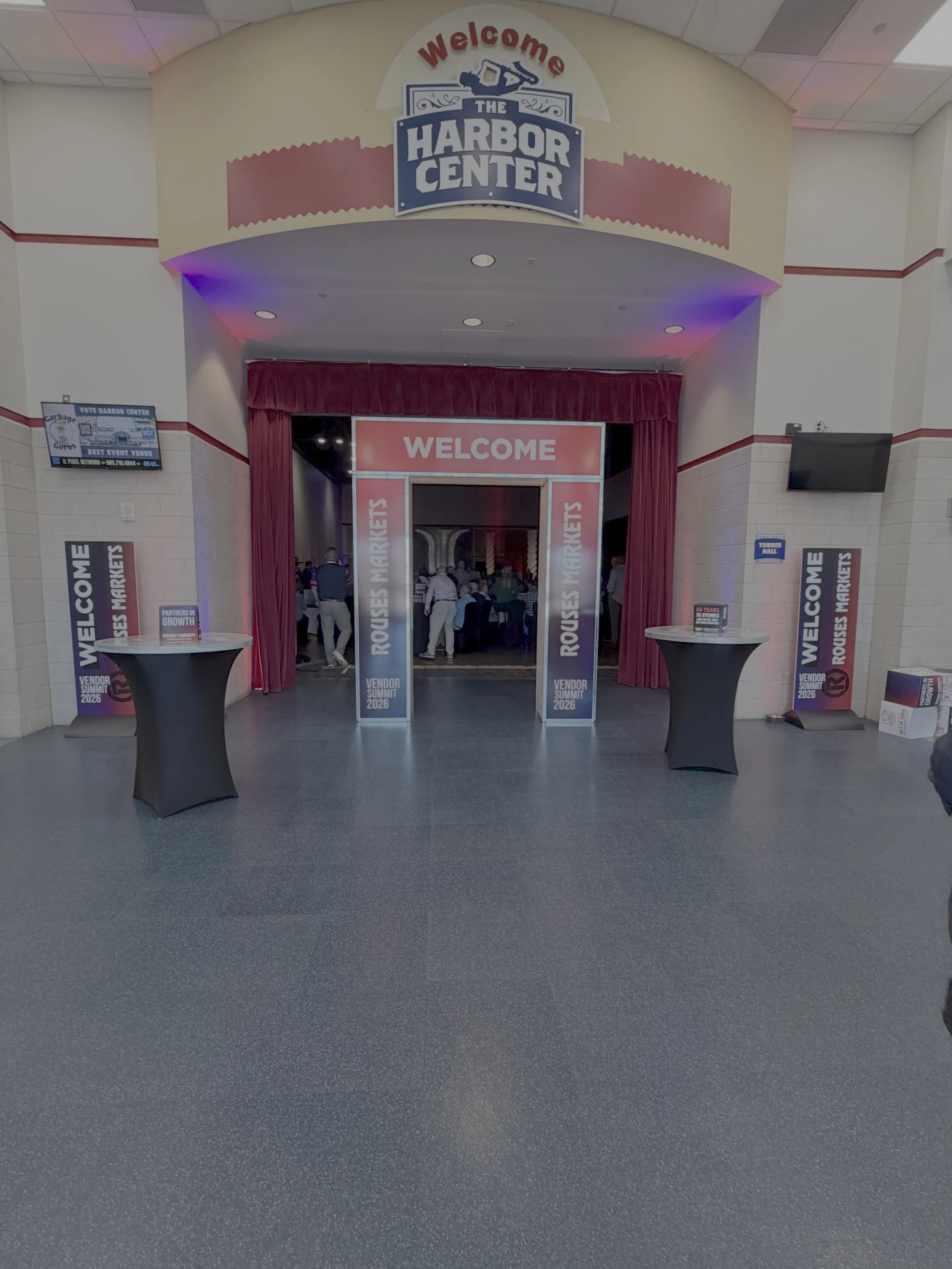 Entrance to the Harbor Center with a welcoming sign, red curtains, and tables for event registration or information. Several people are inside, and banners and screens are visible.