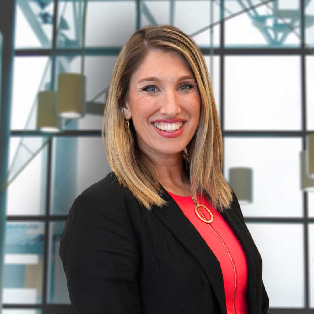 A woman with shoulder-length blonde hair, wearing a black blazer over a red top, smiling in front of a modern glass building.