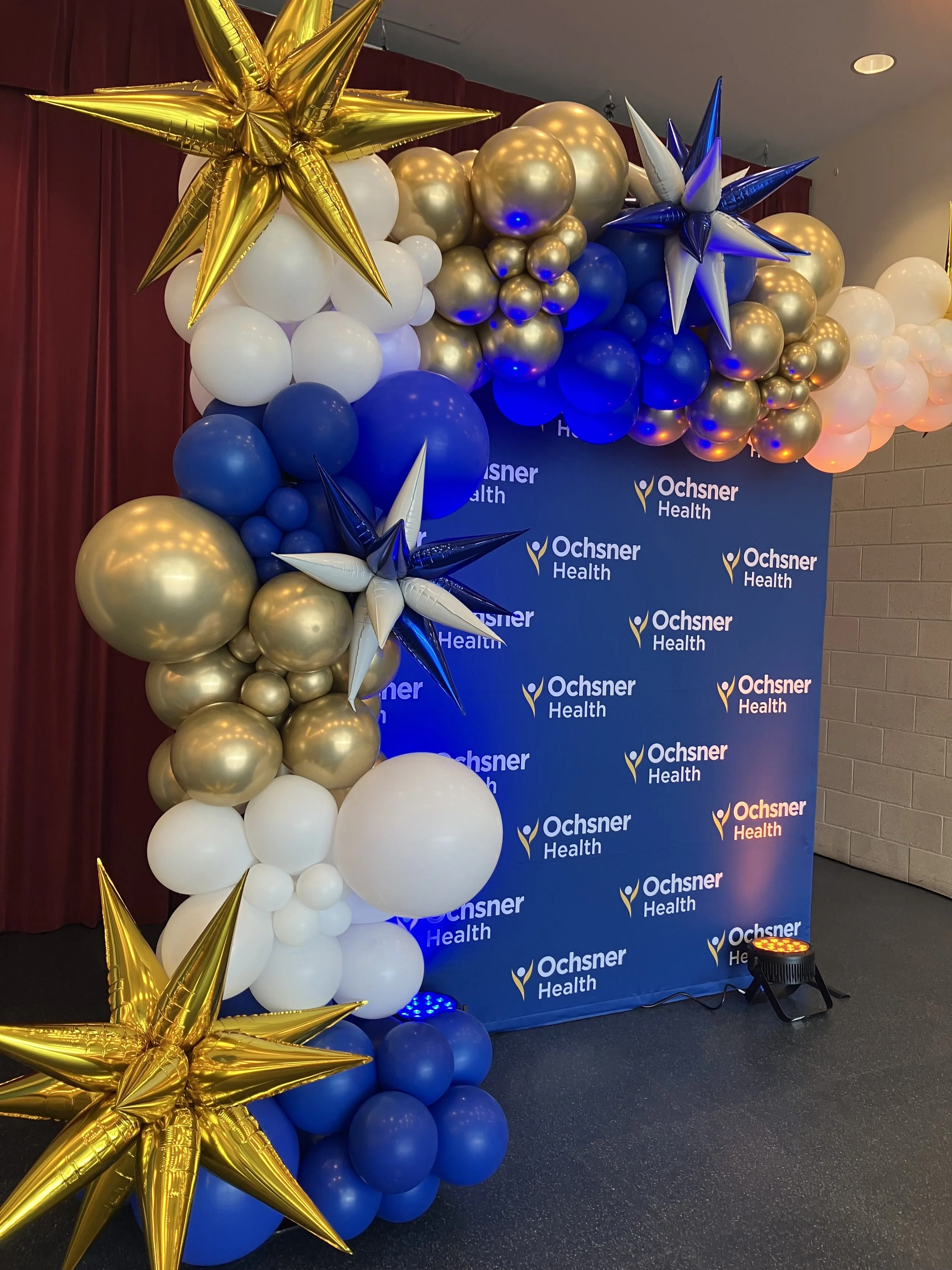 Decorative balloon arrangement with gold, white, and blue balloons, featuring large gold and blue star-shaped balloons, set against a blue backdrop with the Ochsner Health logo.
