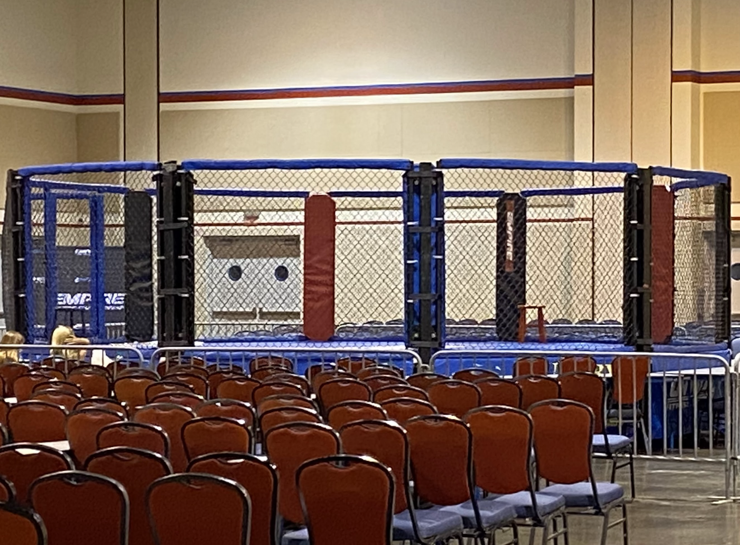 Empty MMA octagon cage with red and blue padding on a stage in a conference room with rows of orange chairs.