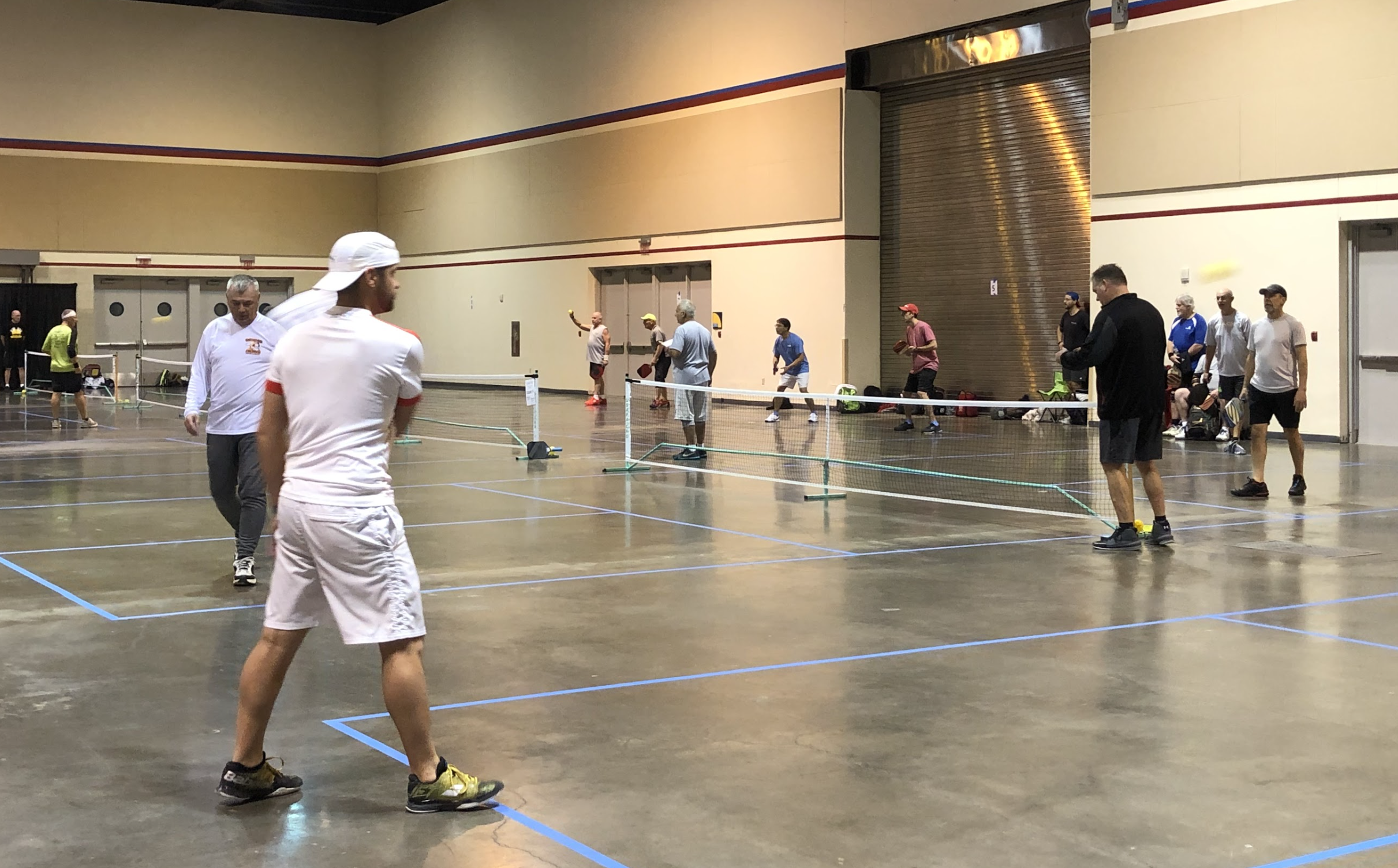 People playing pickleball indoors with nets and paddles, some preparing to serve or hit the ball, in a large gymnasium.
