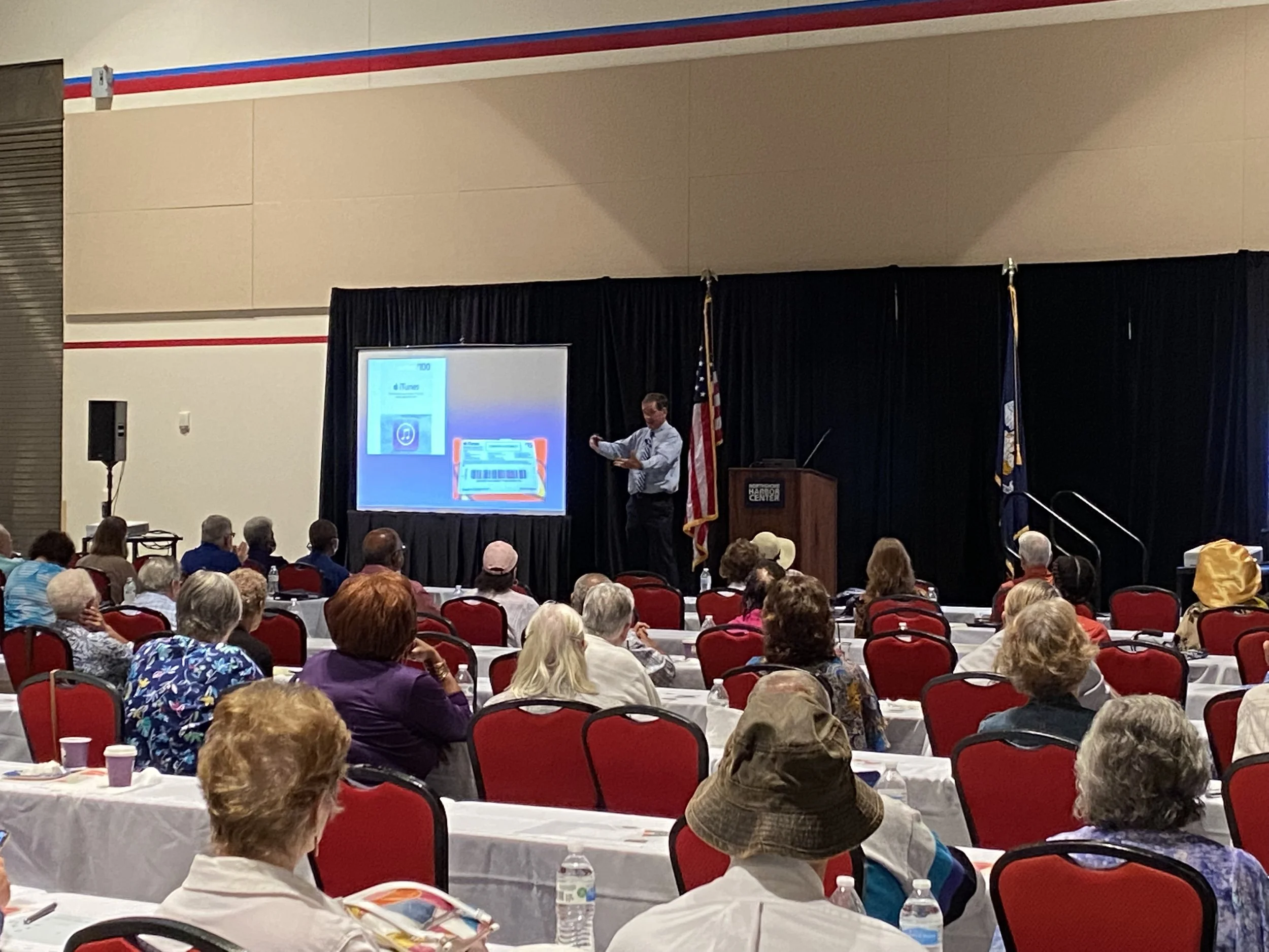 A man giving a presentation on a stage at a conference with a slide about iTunes. The audience is mostly older adults seated at tables with white tablecloths, and flags are displayed behind the presenter.