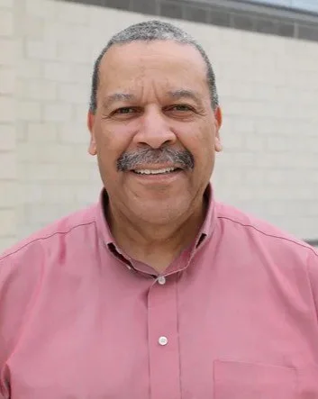 A middle-aged man with short, dark hair and a mustache, smiling at the camera, wearing a pink button-up shirt, standing outdoors in front of a light-colored brick wall.