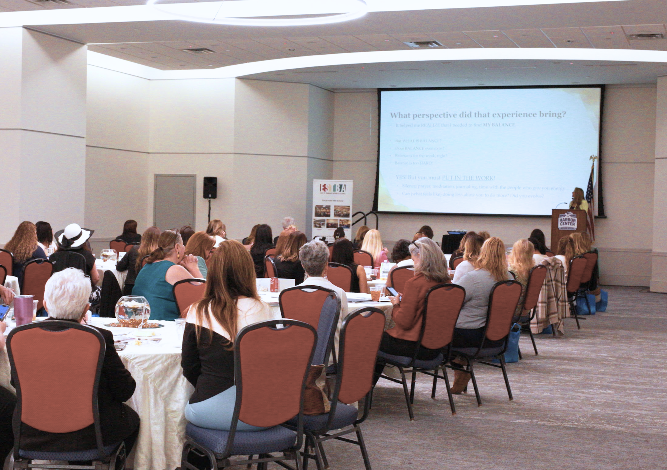Conference room with attendees listening to a presenter at a podium, large screen displaying presentation slides, American flag, and a banner.