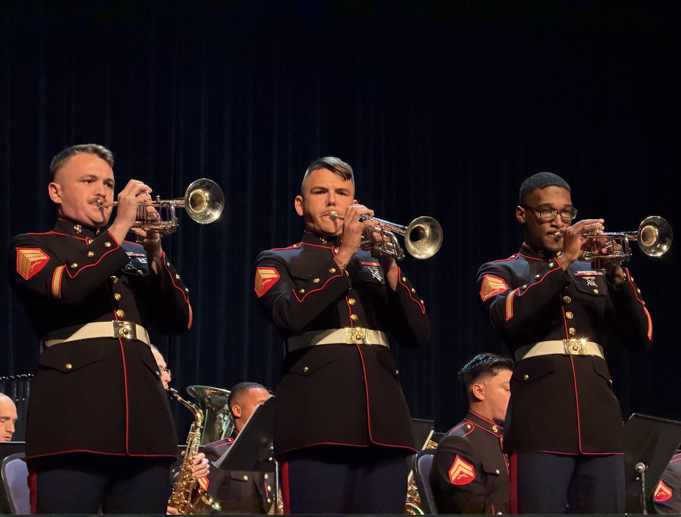Three male military musicians playing trumpets on stage, wearing black uniforms with red and gold accents, in front of a dark curtain.
