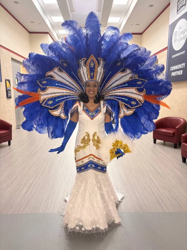 Woman dressed in elaborate costume with a large feathered headdress in blue, orange, and white, in an indoor setting with chairs and signs.