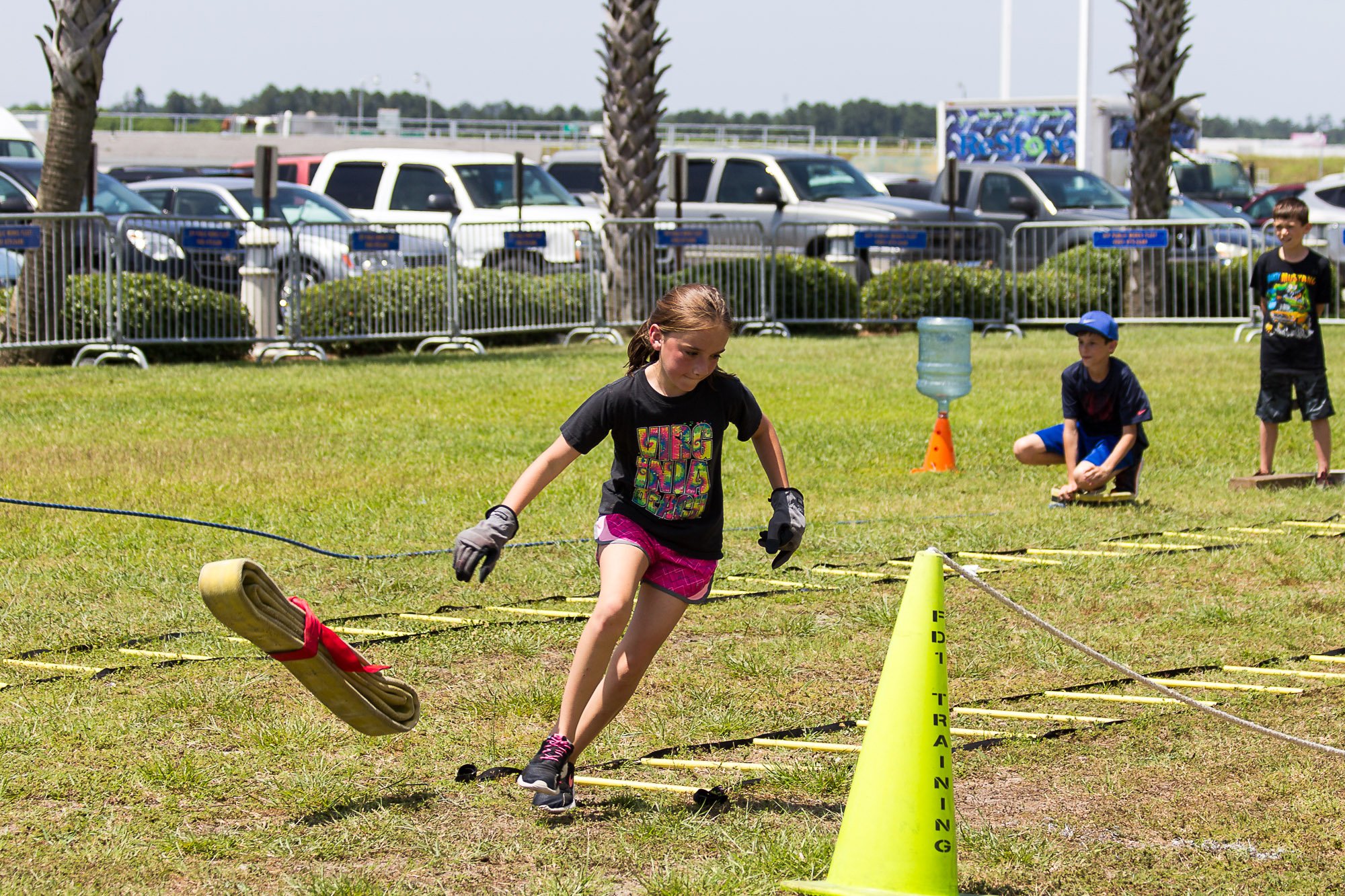 A young girl running in an outdoor obstacle course, with a yellow cone labeled 'Training' and a rolled-up mat attached to a rope. Other children are watching or participating in the background, and parked cars are visible behind a fence, under a sunn