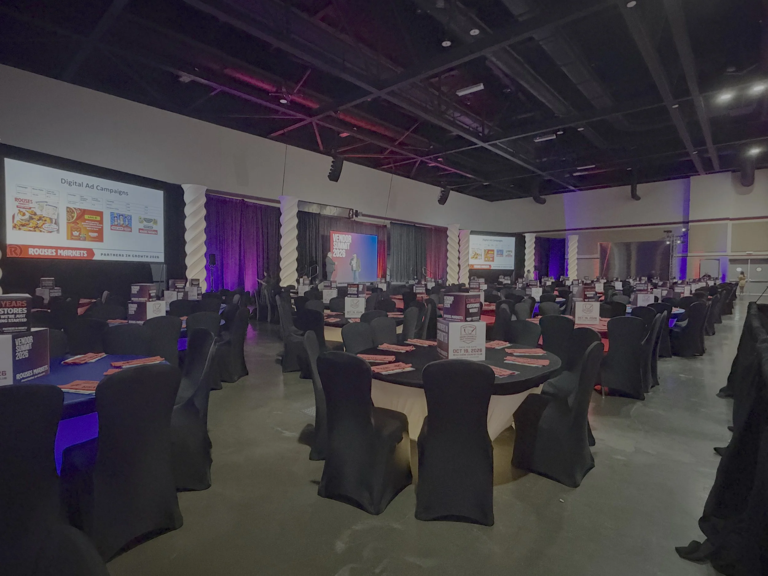 Conference room with rows of chairs, large screens displaying presentation slides, and a stage at the front. The room is dimly lit with purple lighting and signs indicating an event called Vendor Summit 2026.