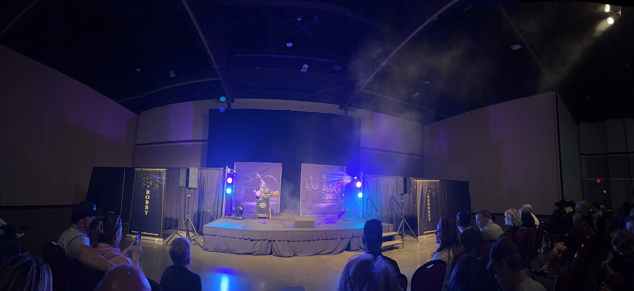 An indoor stage set up for a magic show with colorful lighting, banners advertising 'Robby Bennett', and an audience seated in front.