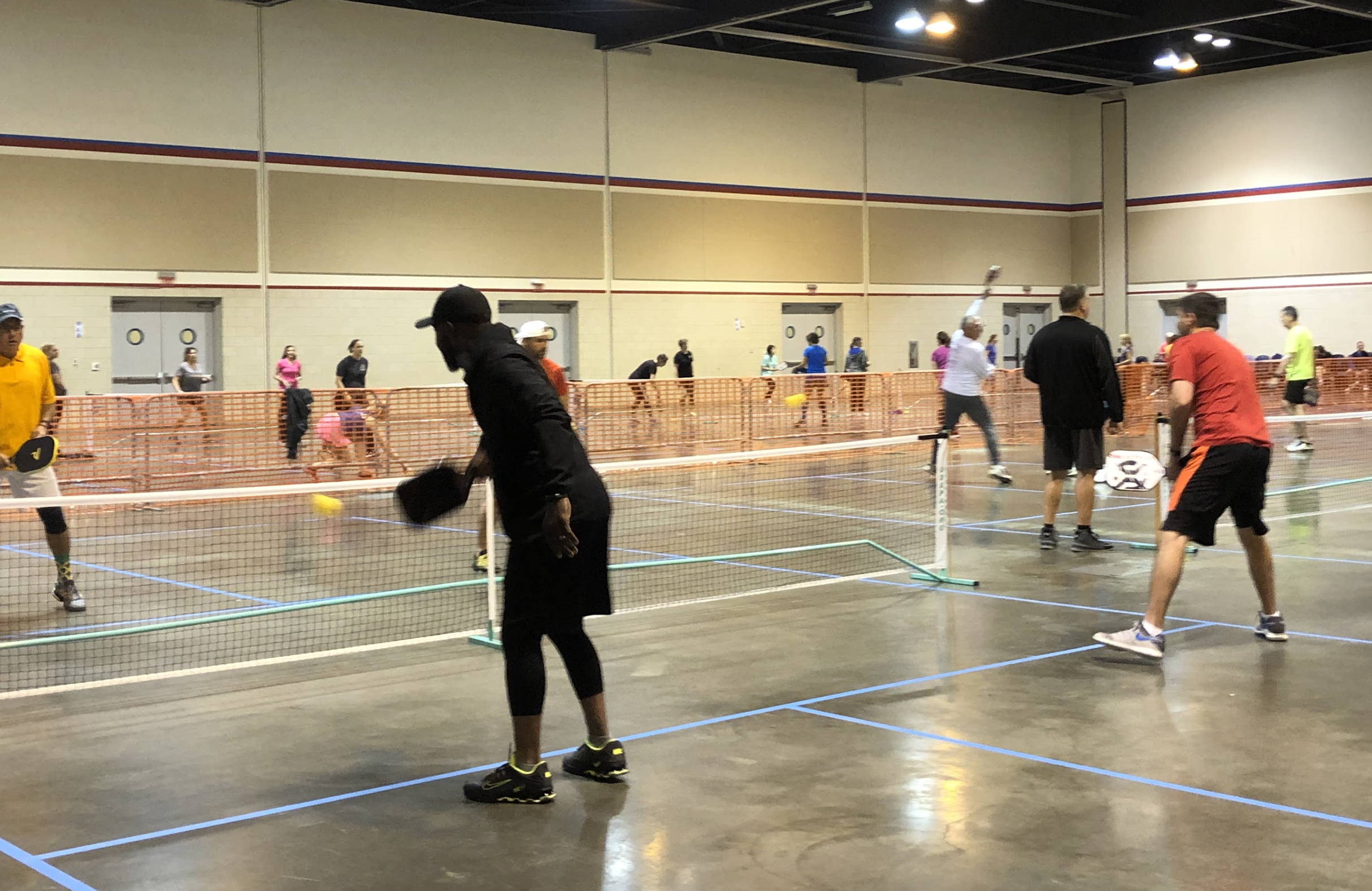 Multiple people playing pickleball in an indoor gym with orange barriers.
