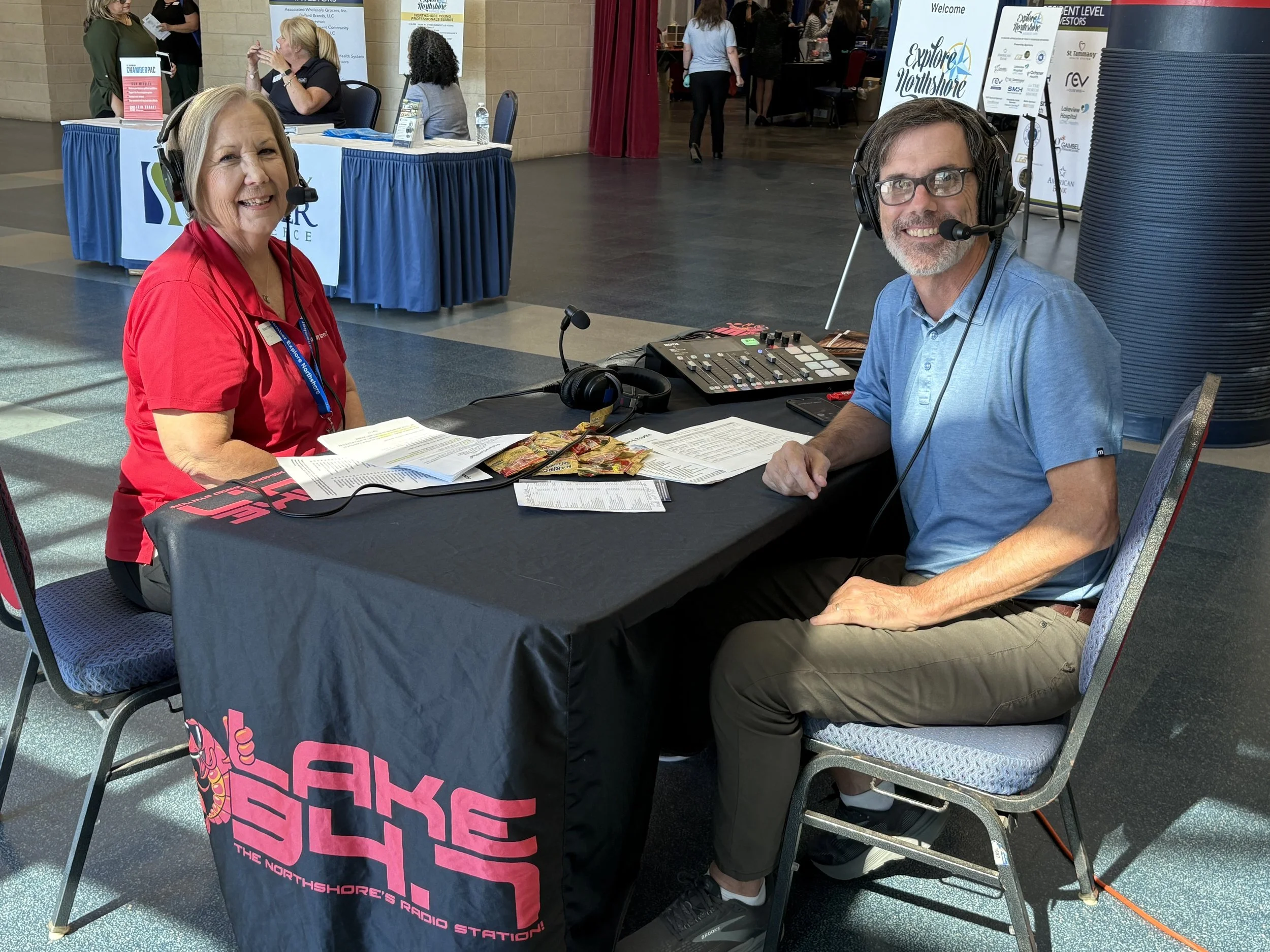 Two radio hosts sitting at a table with broadcasting equipment at an indoor event. The woman on the left is smiling with a headset, wearing a red shirt, and the man on the right is smiling, wearing glasses and a blue polo shirt. The table has a black