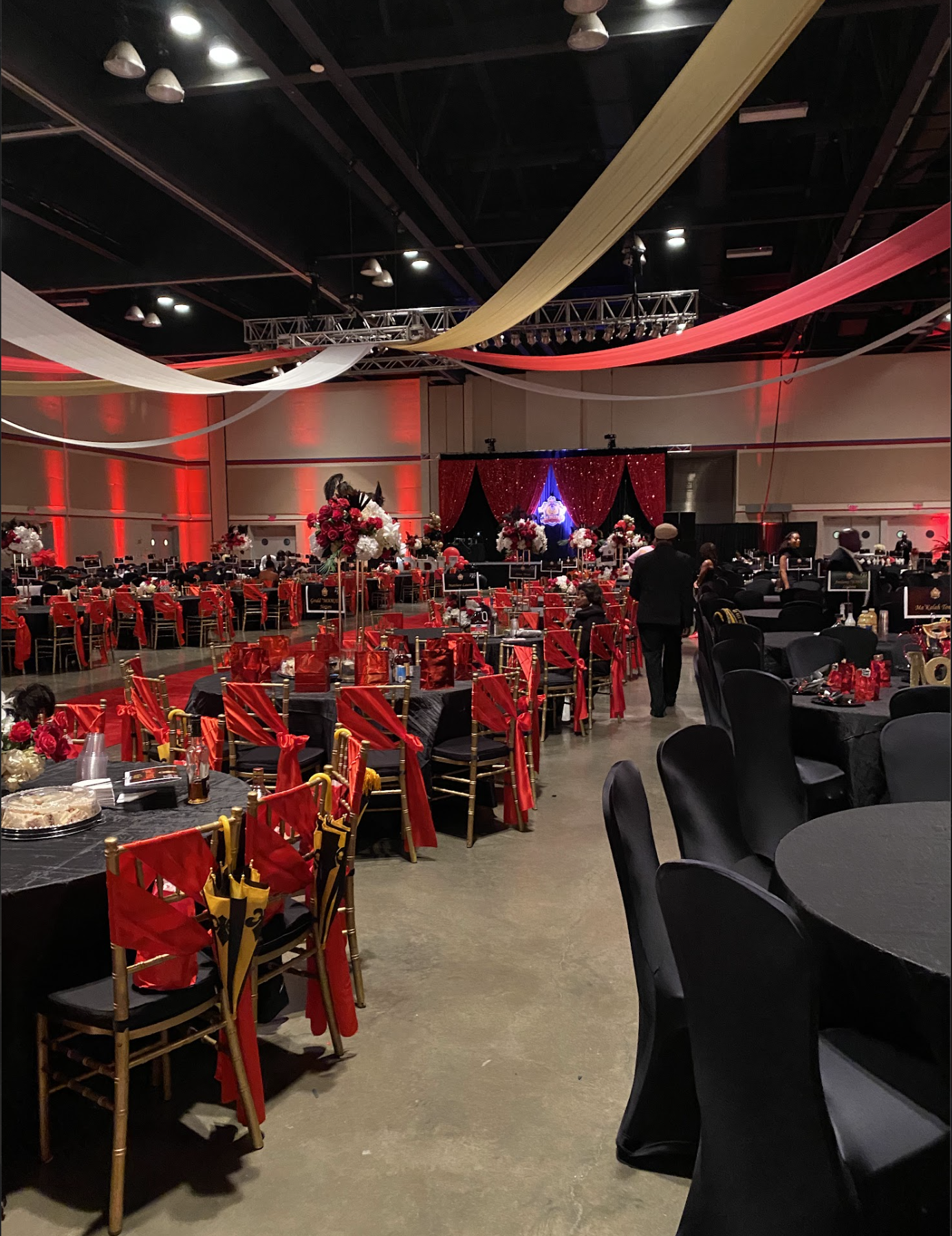 Elegant event hall decorated with red, black, gold, and white drapes hanging from the ceiling, floral centerpieces with roses and carnations, tables set with black tablecloths, red sashes, and gift boxes, and a stage with a red curtain backdrop.