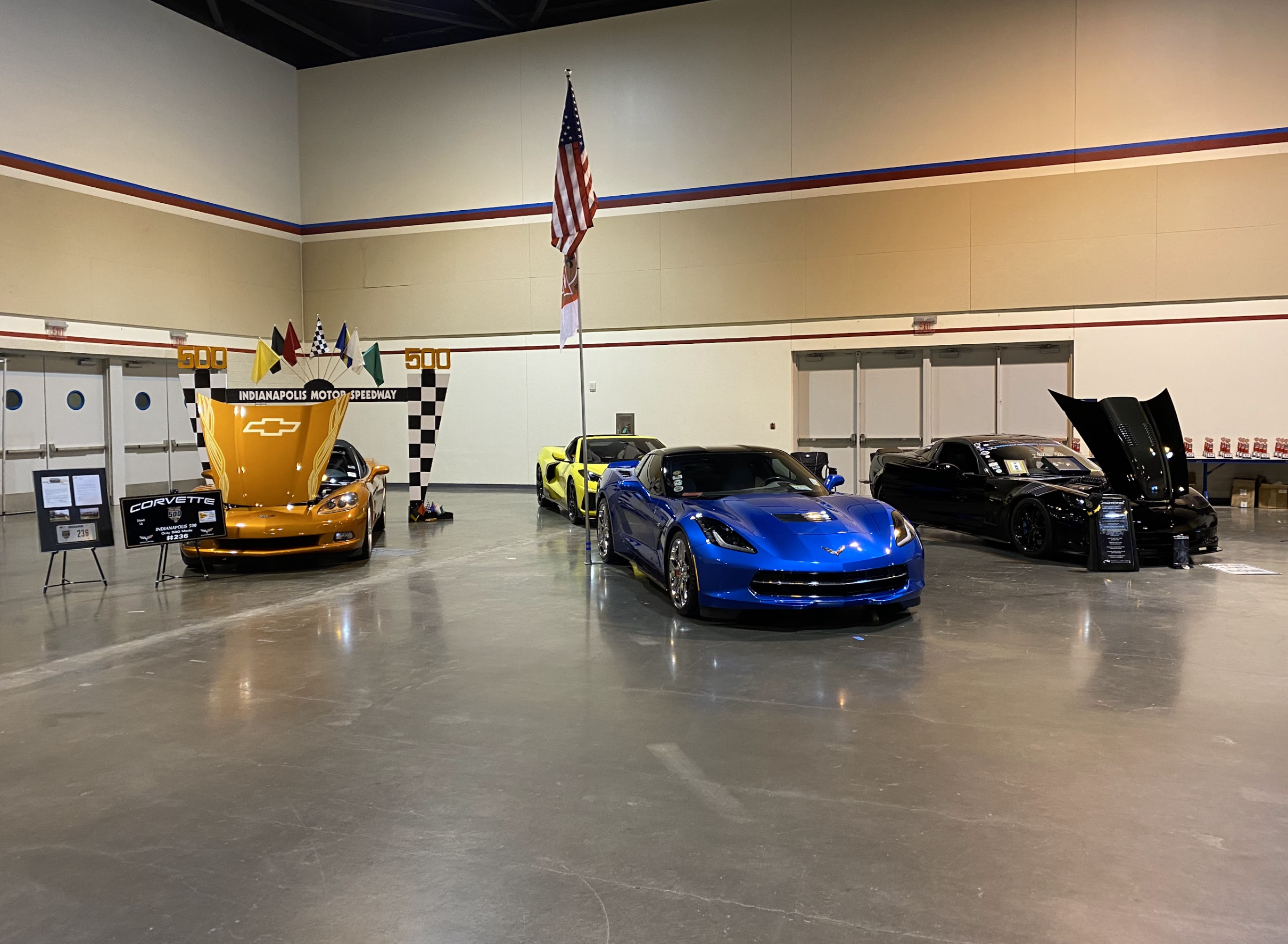 Four Chevrolet sports cars on display at Indianapolis Motor Speedway, with a yellow car on a platform with flags, a blue car in the foreground, a black car with its hood open, and a yellow and black car in the background. American flag and Indianapol