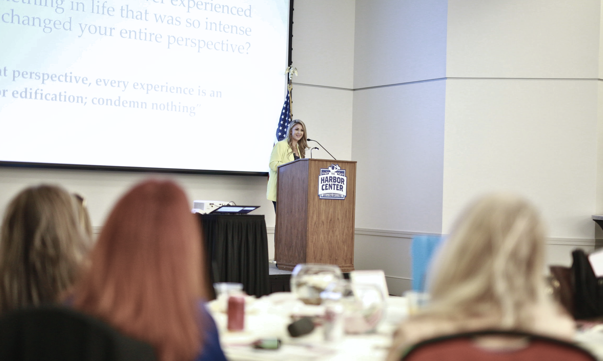 A woman speaking at a podium with a sign that says 'Harbor Center' in a conference room, with an American flag behind her and audience members in front.