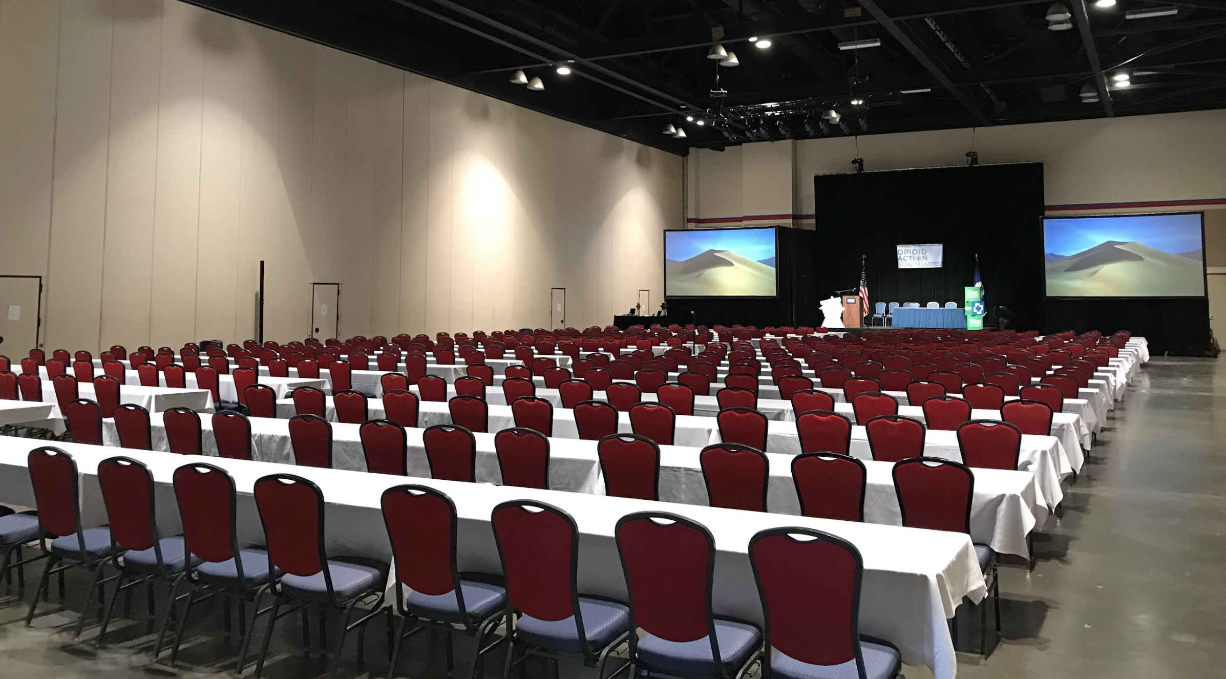 Empty conference hall with rows of red chairs and long tables covered with white tablecloths, facing a stage with two large screens displaying desert dunes, a podium, American and state flags, and a banner reading 'opioid action summit'.