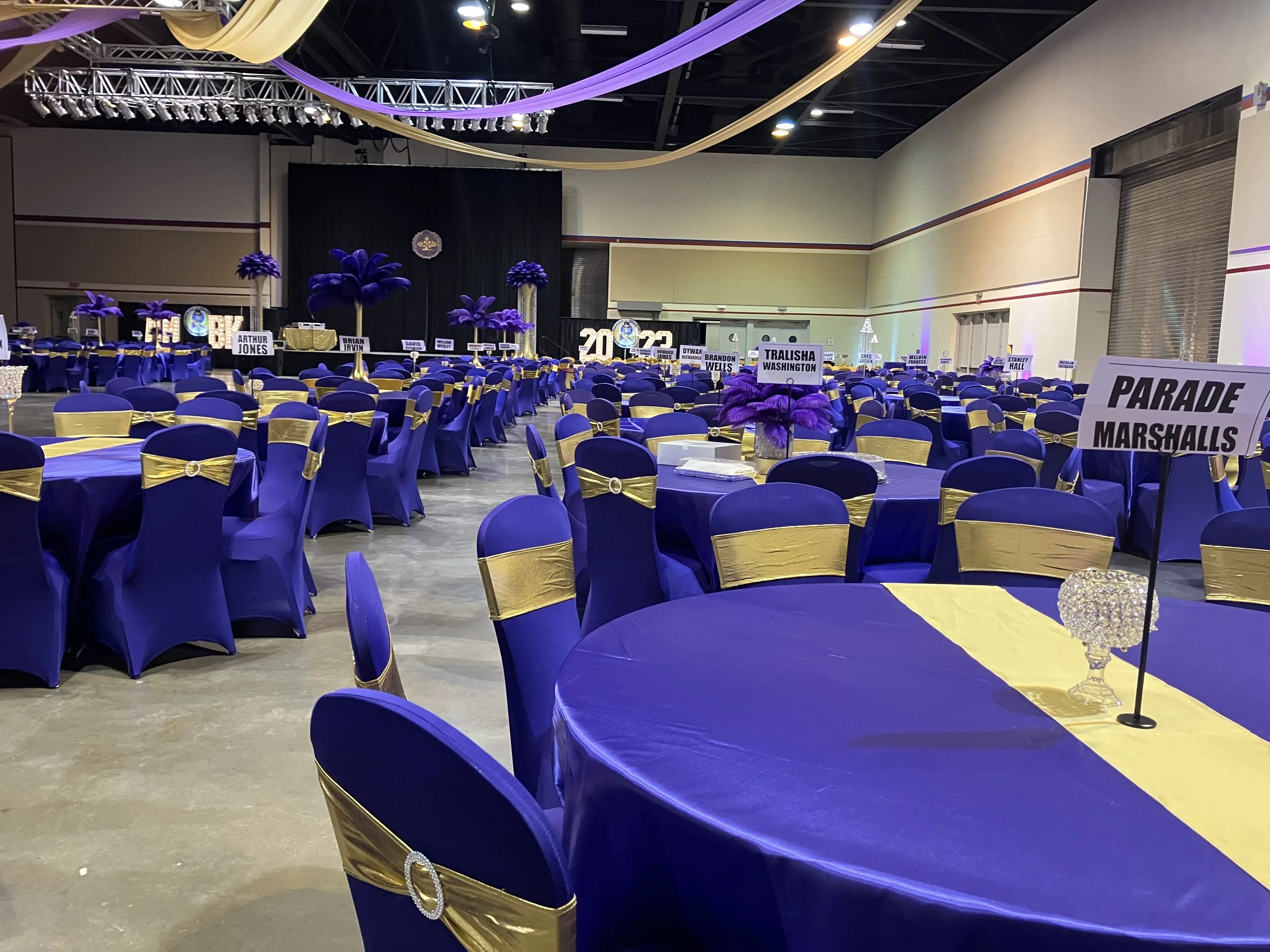 A large banquet hall decorated in purple and gold for a graduation celebration, featuring multiple round tables with purple tablecloths, gold sashes on the chairs, and purple feather centerpieces. There are signs indicating different graduation class